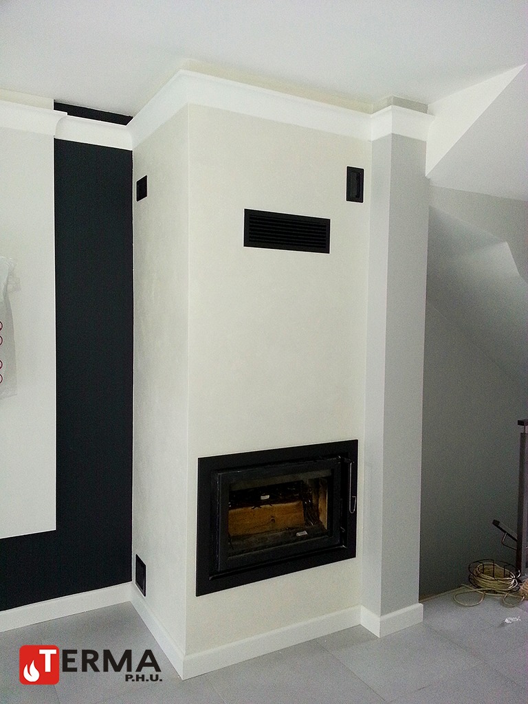 Biały, zabudowany wkład z czarną ramą i kratkami wentylacyjnymi w nowoczesnym salonie z szarą podłogą i czarną ścianą akcentującą.