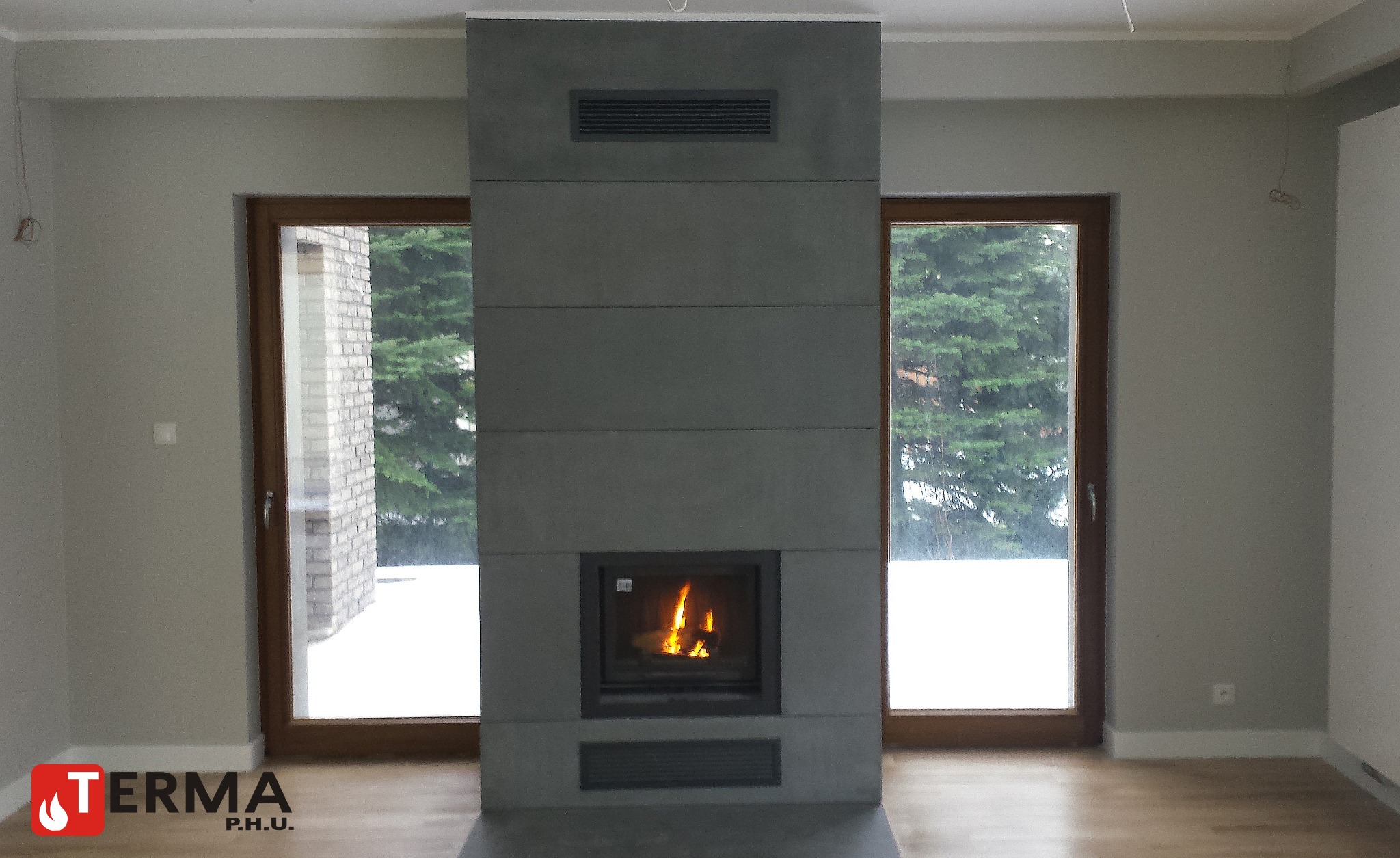 Nowoczesny, minimalistyczny wkład kominkowy z szarej obudowie z poziomymi panelami, płomień widoczny przez szybę, jasne wnętrze z dużymi oknami.