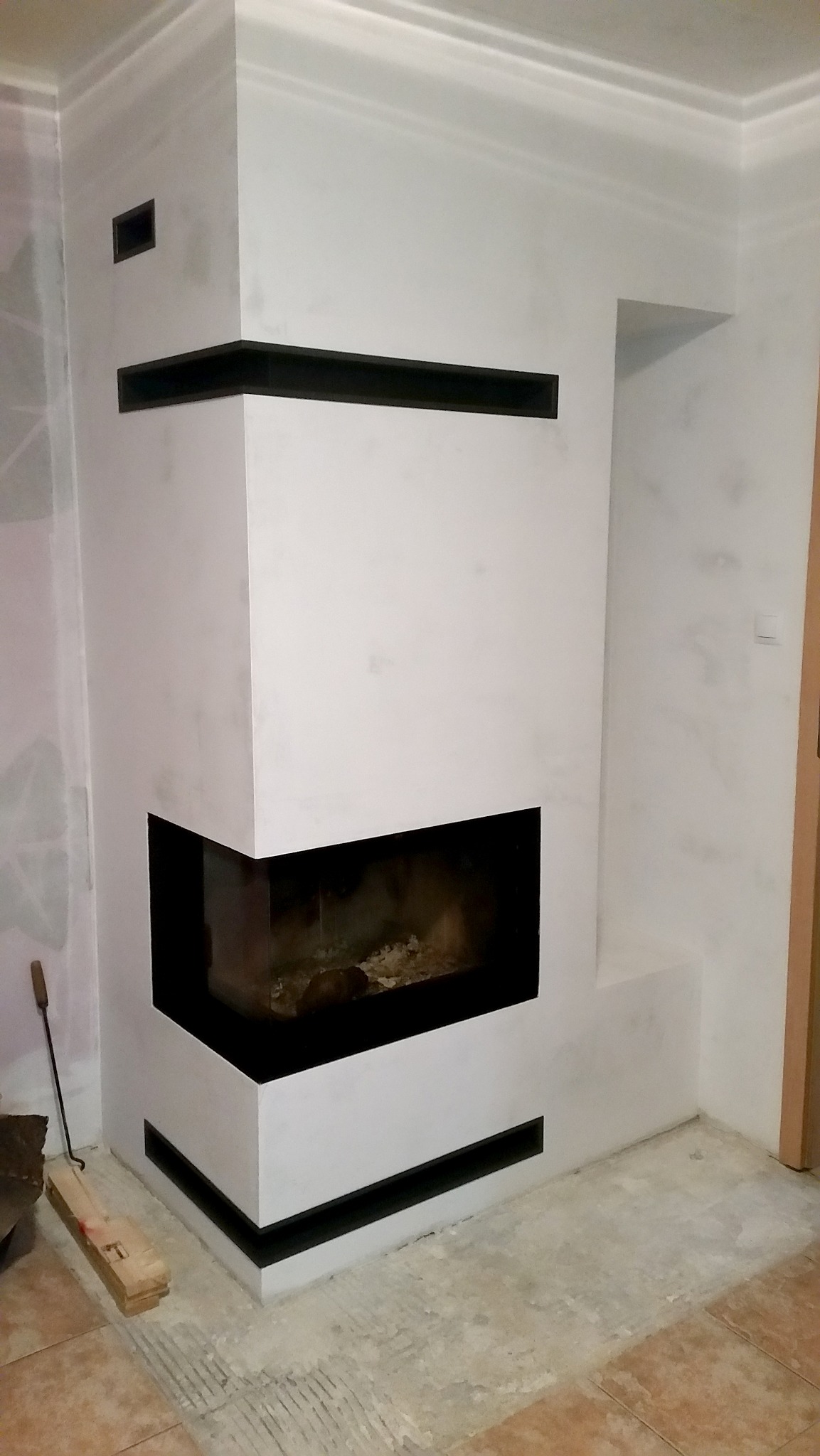 Biały narożny wkład kominkowy z czarnymi akcentami, widoczny w trakcie wykańczania, z narzędziami obok i niedokończoną podłogą.