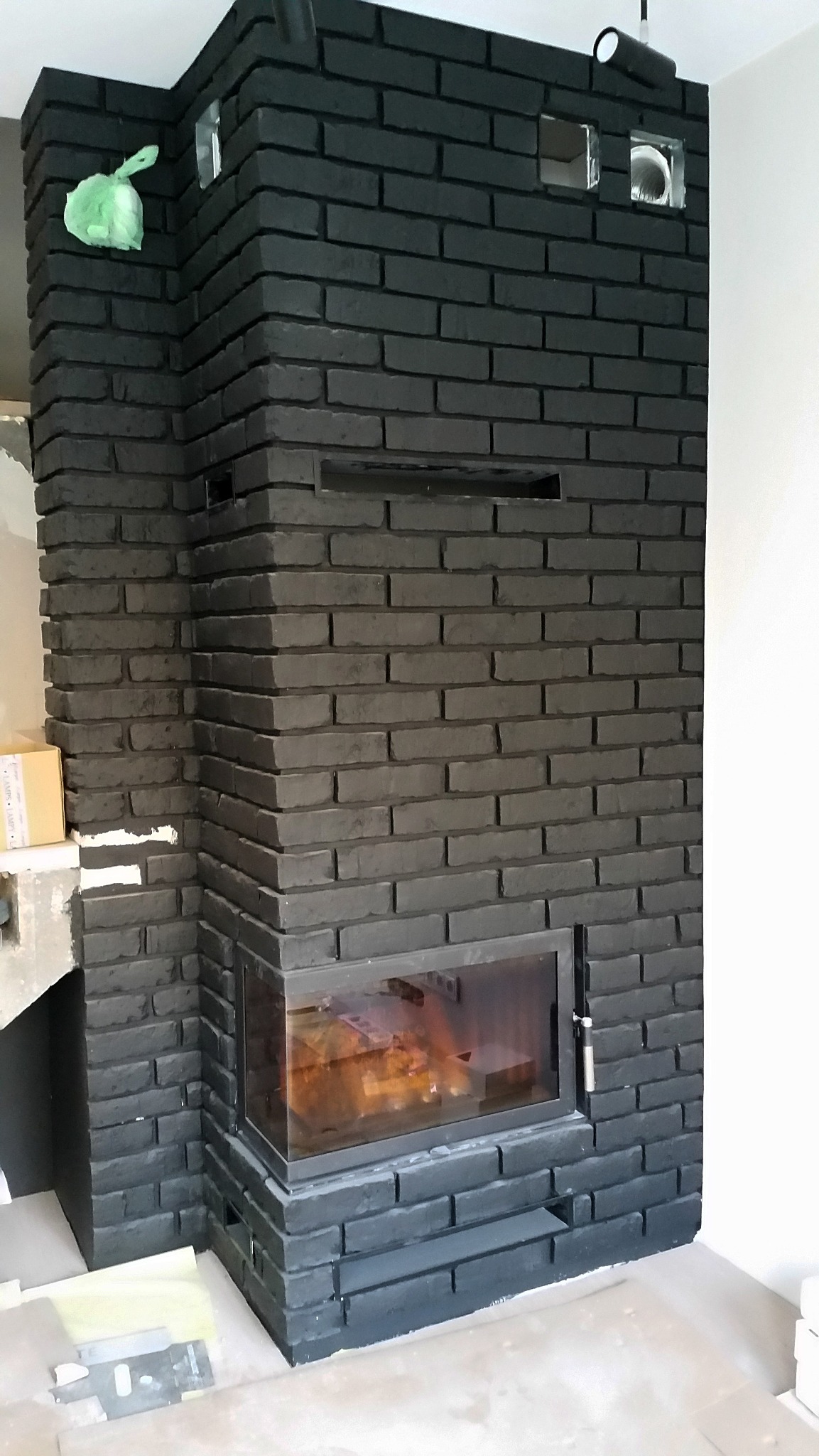 Nowoczesny, czarny piec obłożony cegłą, z widocznym paleniskiem i elementami instalacji wentylacyjnej w trakcie montażu.