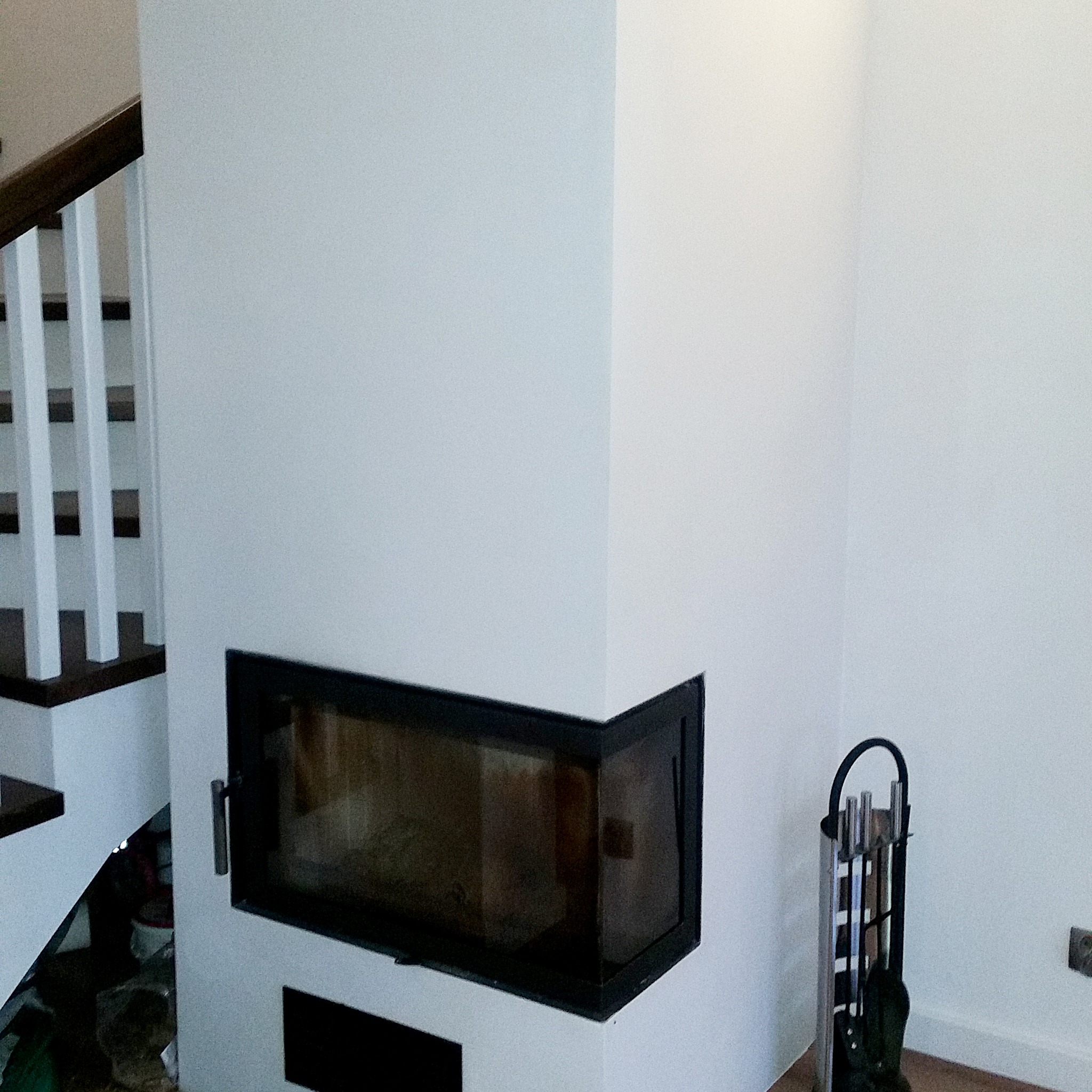 Narożny kominek z czarną ramą, widok z boku, obok schody z drewnianymi elementami i zestaw akcesoriów kominkowych ze szczypcami, pogrzebaczem i zmiotką.