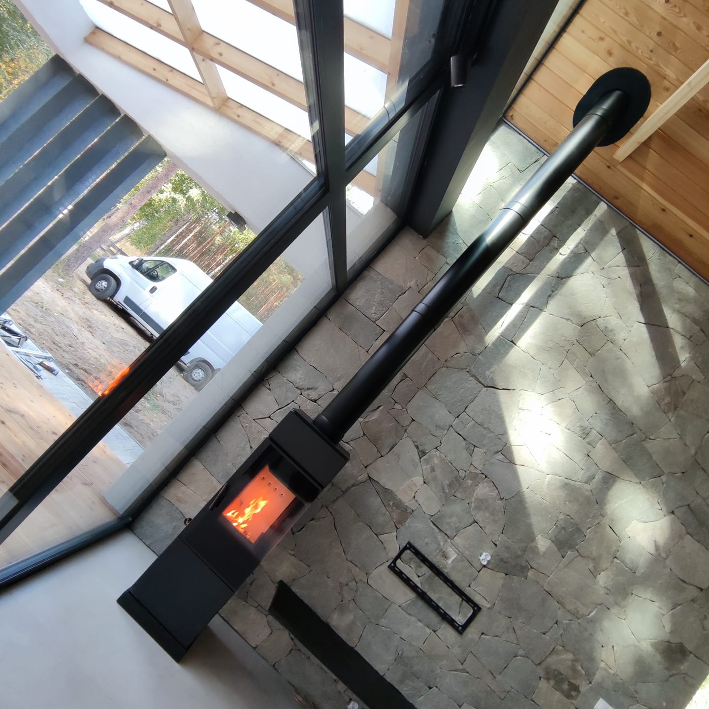 Nowoczesny, czarny piec wolnostojący z płomieniami w palenisku, widoczny z góry na tle kamiennej ściany i podłogi, z oknem wychodzącym na zewnątrz, gdzie stoi biały samochód dostawczy.