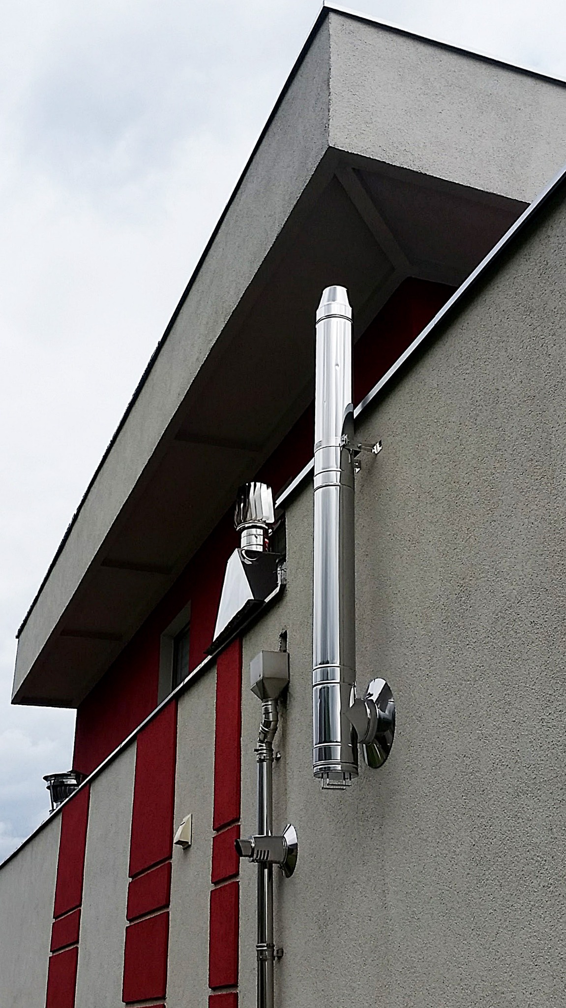 Zewnętrzny, stalowy system odprowadzania spalin z kotła grzewczego, zamontowany na elewacji budynku z czerwono-białymi pasami.
