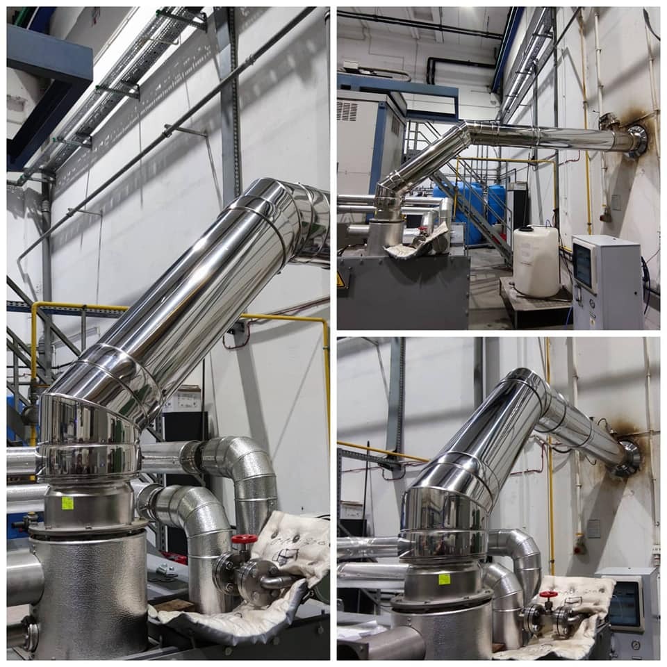 Montaż systemu odprowadzania spalin z izolacją termiczną, widoczny fragment instalacji z zaworem i rurami ze stali nierdzewnej w hali przemysłowej.