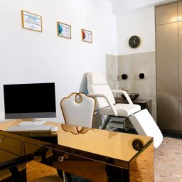 Justyna Kafel-Wituszyńska - Wnętrze gabinetu z białym fotelem zabiegowym, biurkiem z lustrzanym blatem, komputerem, oraz ścianą udekorowaną certyfikatami w złotych ramkach.