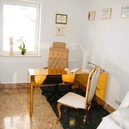 Justyna Kafel-Wituszyńska - Jasne wnętrze gabinetu z biurkiem w kolorze złota, białym krzesłem i dyplomami na ścianie, widok na okno z rośliną i figurką.