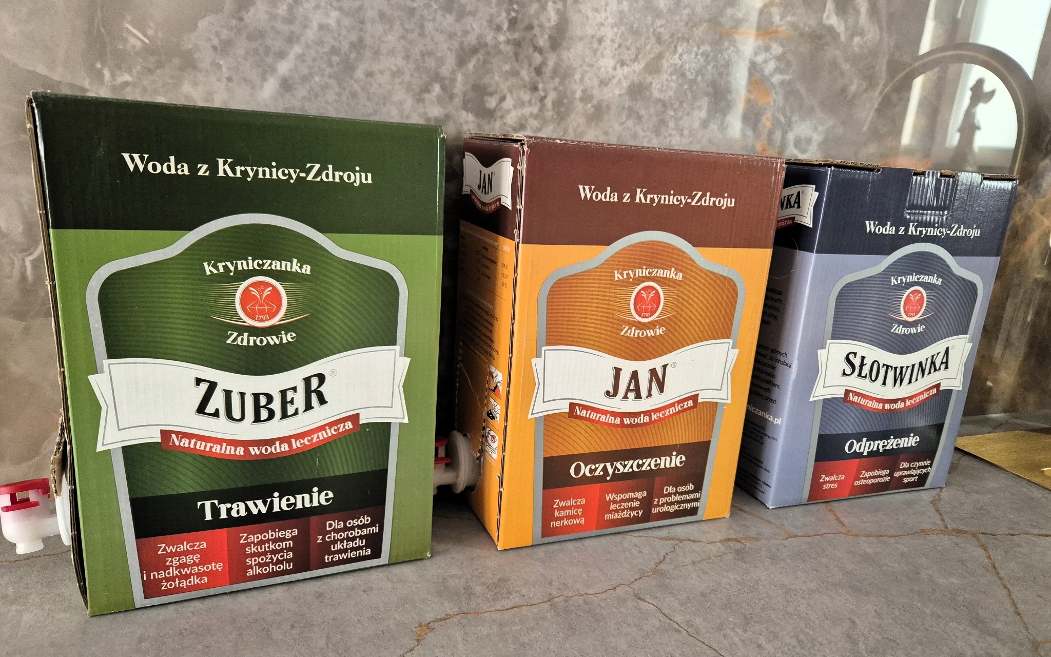 Trzy kartonowe opakowania wody leczniczej Kryniczanka: Zuber, Jan i Słotwinka, ustawione obok siebie na marmurowym blacie, z widocznymi kranikami do nalewania.