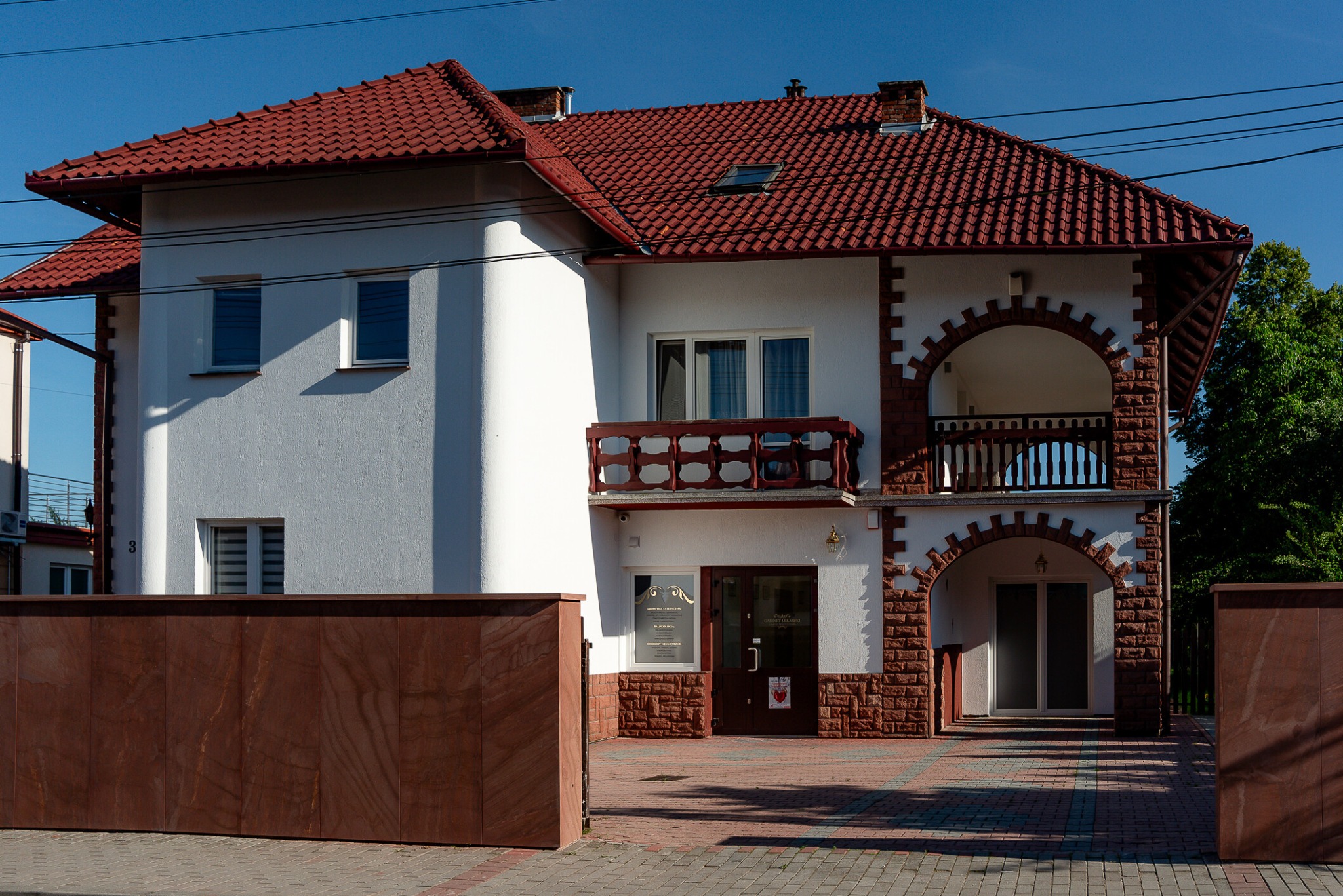 Dwupiętrowy budynek z czerwonym dachem i ceglanymi elementami dekoracyjnymi, widoczny szyld z napisem 'Medycyna Estetyczna' na oknie obok wejścia.