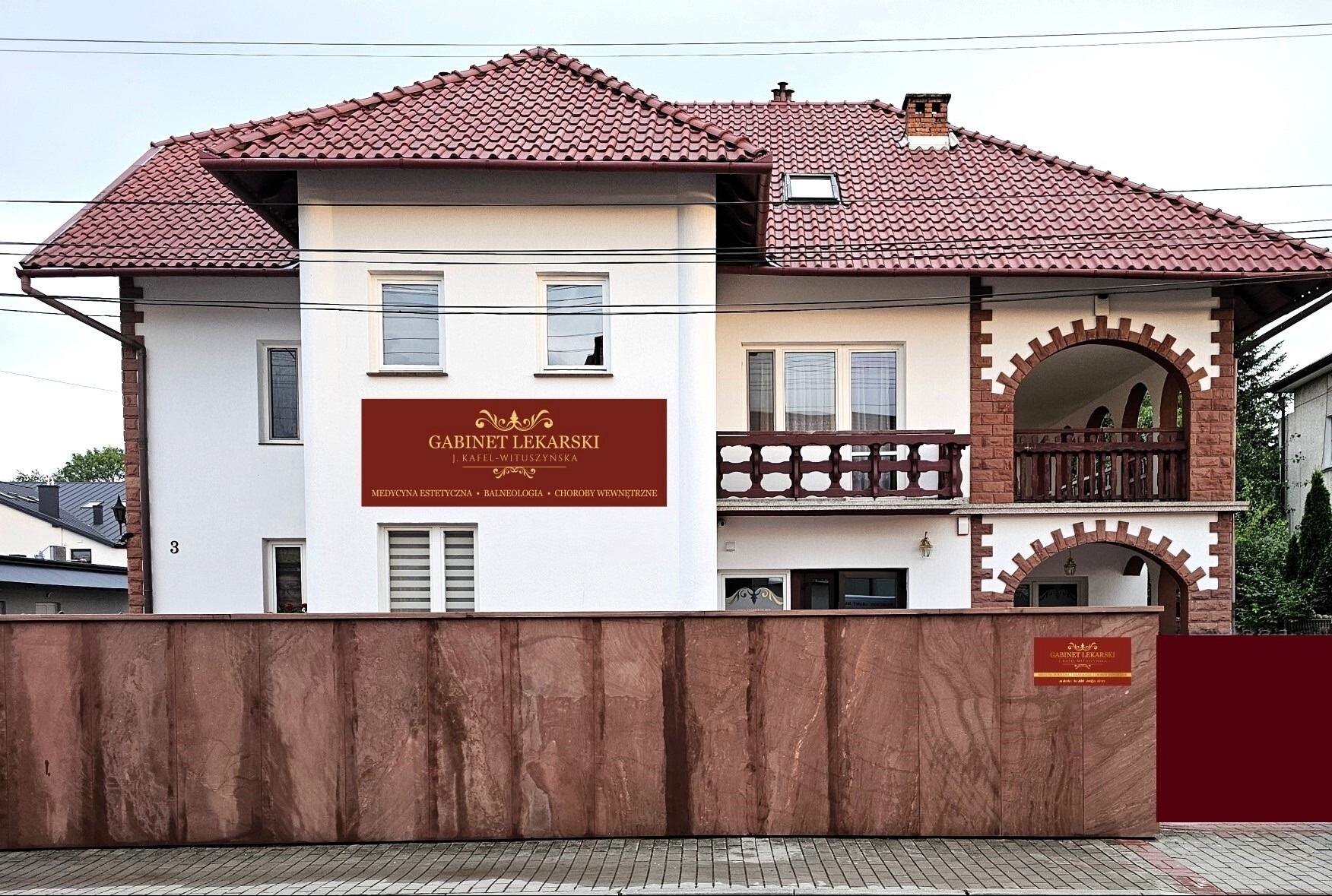 Fasada dwupiętrowego budynku z czerwonym dachem, tablicą informacyjną 'Gabinet Lekarski' z ofertą medycyny estetycznej, balneologii i chorób wewnętrznych, oraz ogrodzeniem z kamienia.