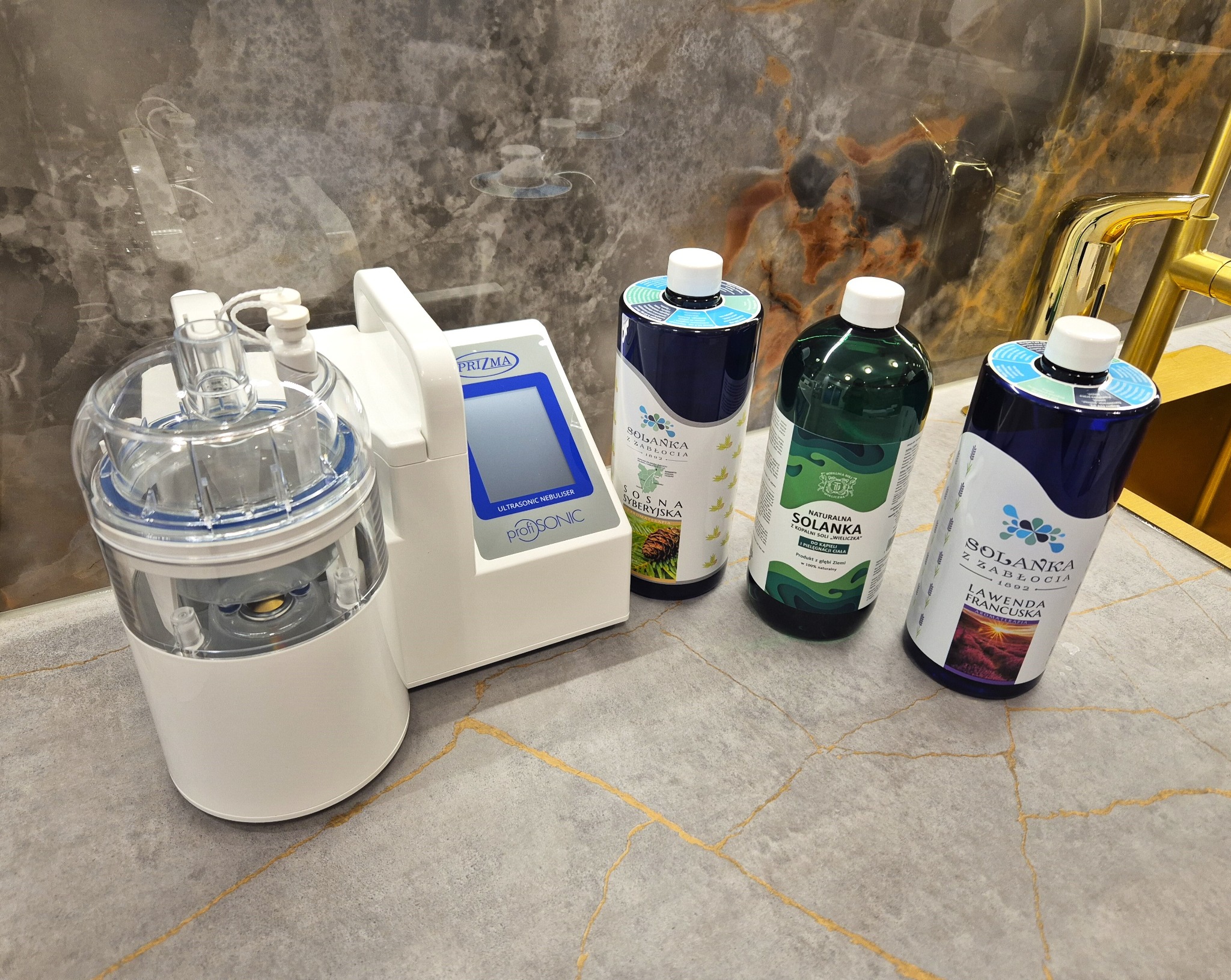 Ultradźwiękowy nebulizator PriZma ProfiSONIC obok trzech butelek solanki z Zabłocia o różnych zapachach: sosna syberyjska, naturalna solanka i lawenda francuska, na blacie z szarymi żyłkami.