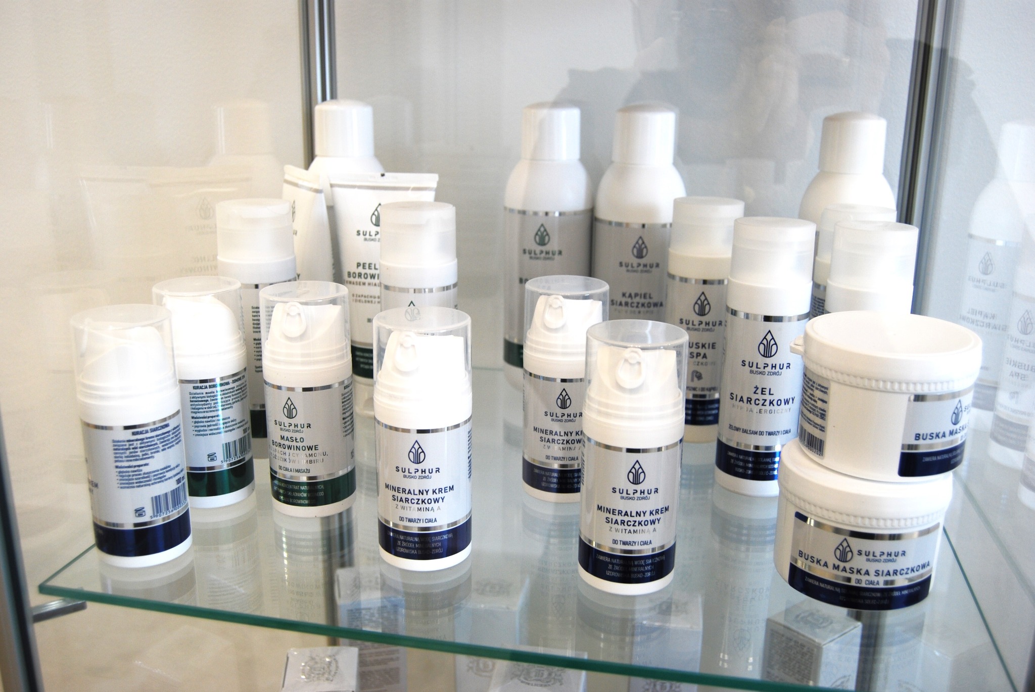Półka z produktami Sulphur Busko Zdrój: kremy, maski, peelingi i żele siarczkowe w białych opakowaniach z srebrnymi etykietami, eksponowane w szklanej gablocie.