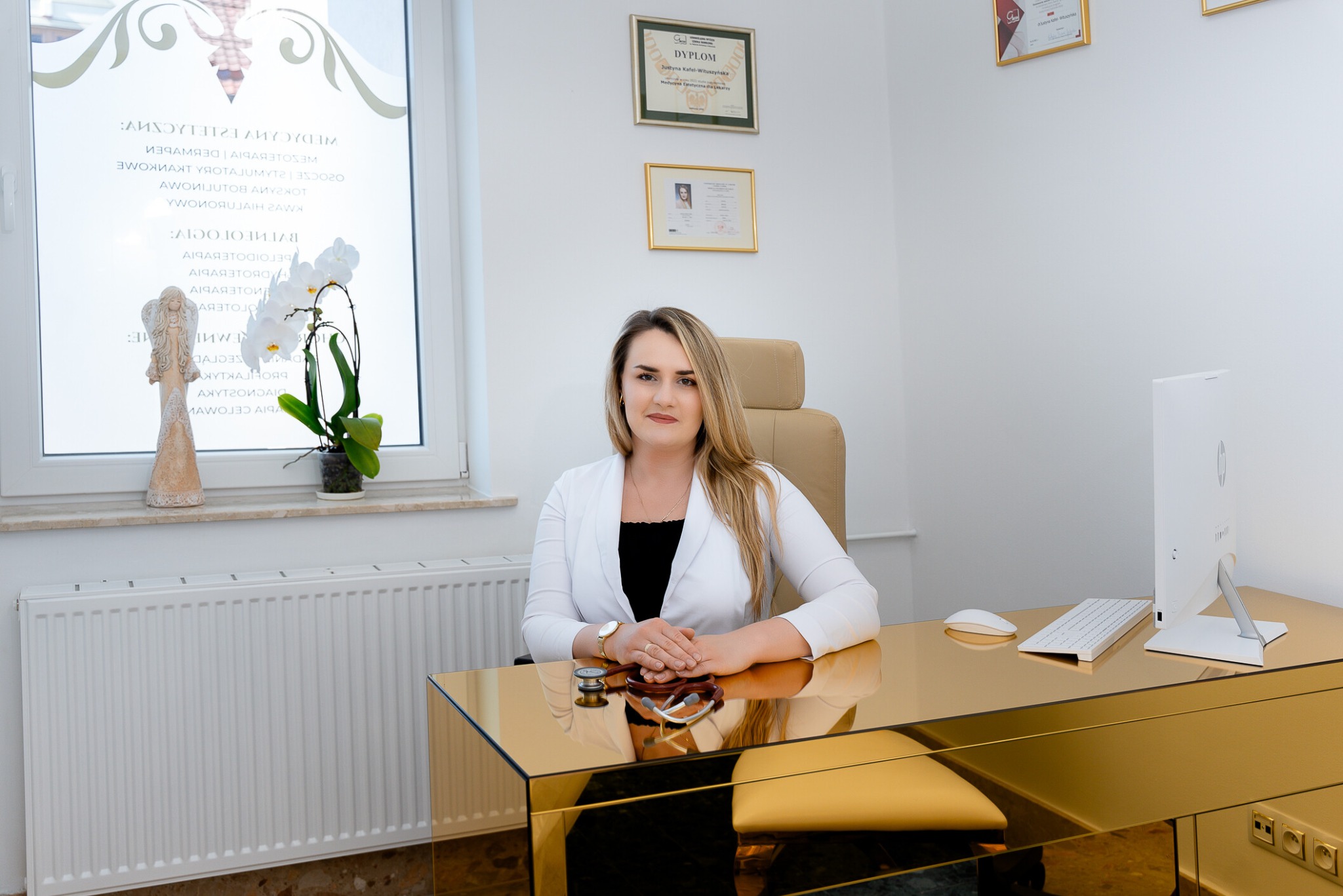 Lekarz medycyny estetycznej w białym fartuchu siedzi za złotym biurkiem w jasnym gabinecie, na biurku stetoskop, w tle okno z dekoracyjną naklejką i kwiatami, na ścianie dyplomy.