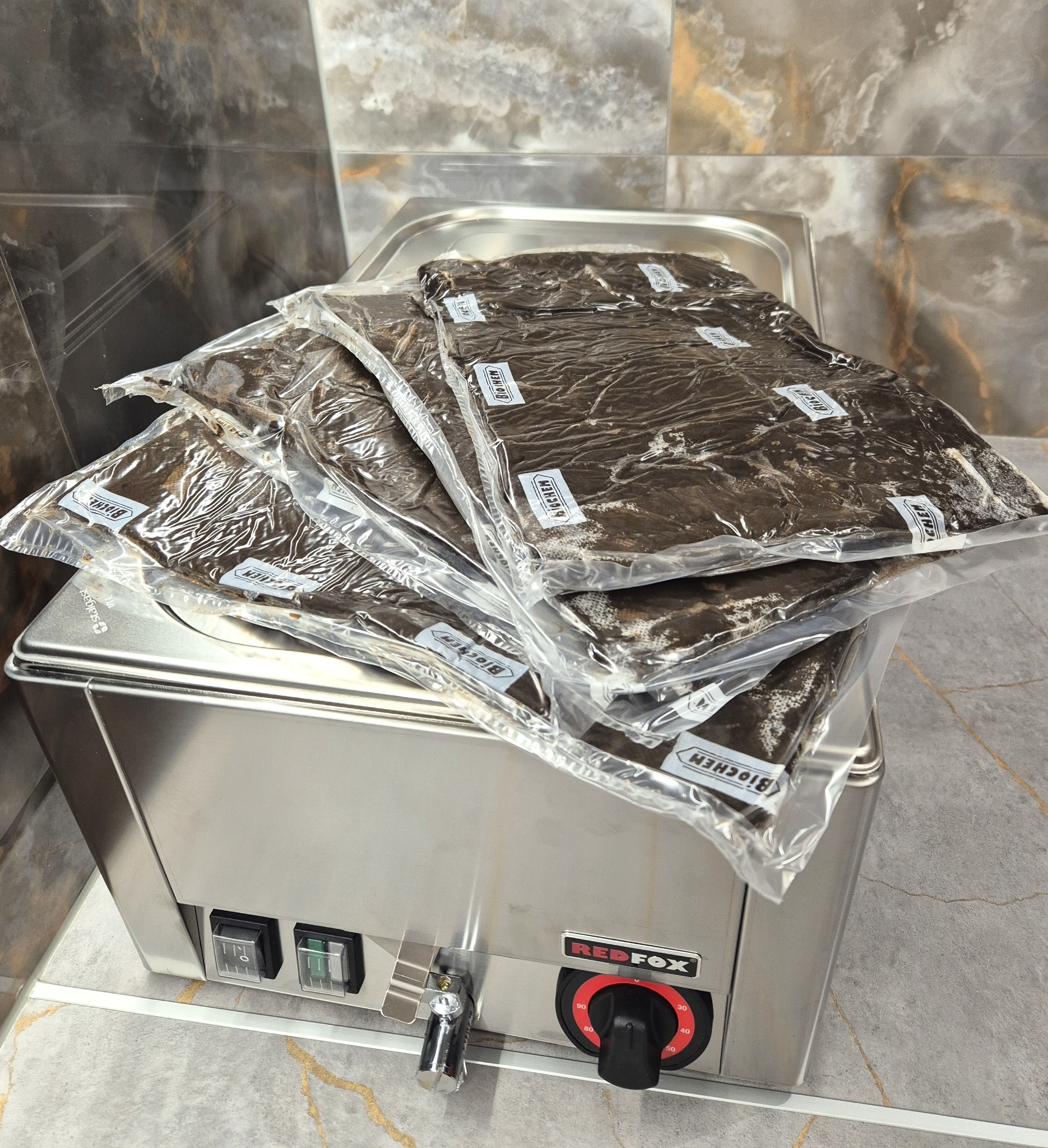 Pakiety z brązową zawartością, opatrzone etykietami BIOCHEM, ułożone na metalowej maszynie marki REDFOX z pokrętłem regulacji temperatury, na tle marmurowych płytek.