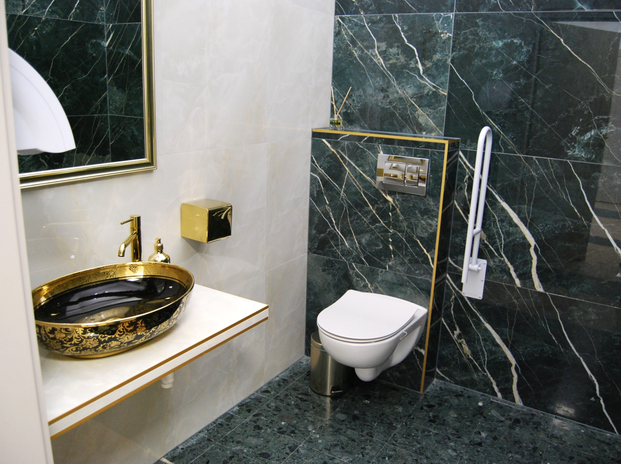 Luksusowa łazienka z czarno-złotą umywalką nablatową, marmurowe płytki w odcieniach zieleni i bieli, wisząca toaleta i złote akcenty.