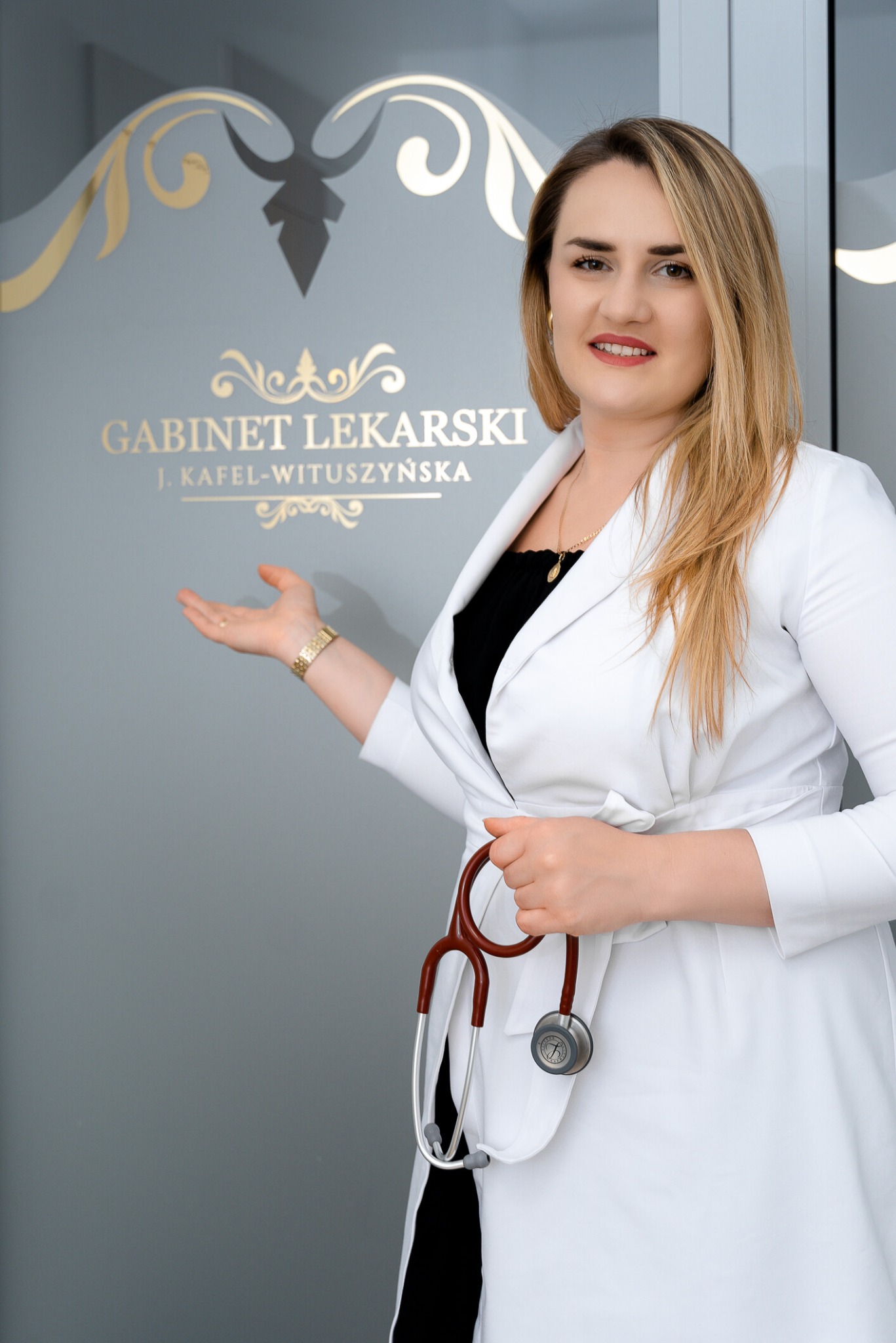 Uśmiechnięta lekarka w białym fartuchu, z brązowym stetoskopem w dłoni, prezentuje gabinet lekarski z eleganckim złotym napisem na szarej ścianie.