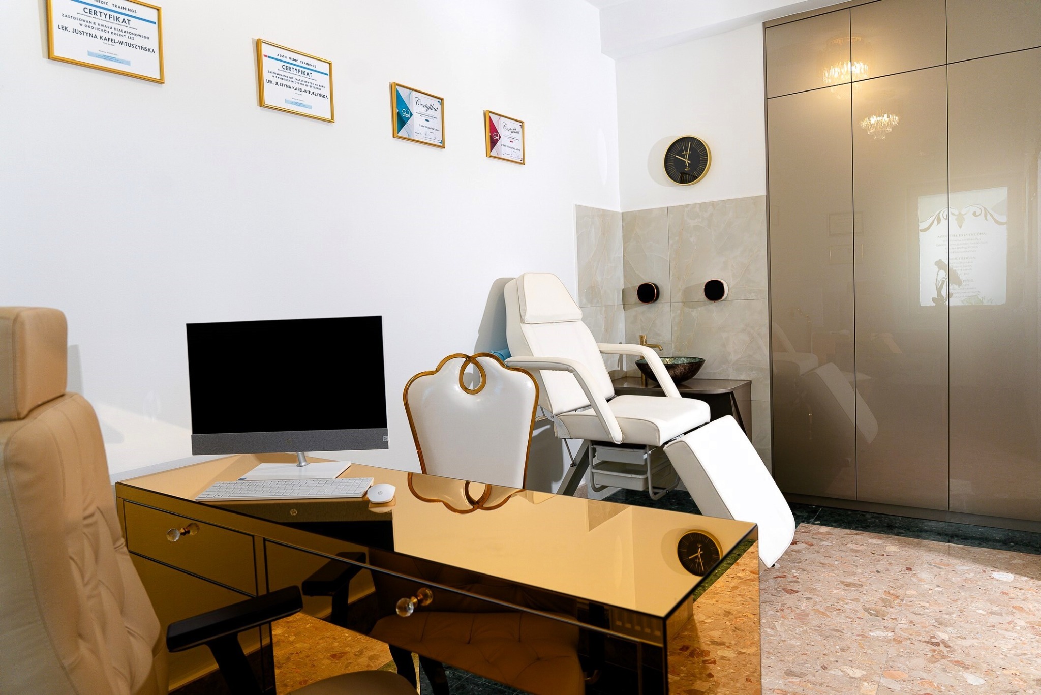 Wnętrze gabinetu z białym fotelem zabiegowym, biurkiem z lustrzanym blatem, komputerem, oraz ścianą udekorowaną certyfikatami w złotych ramkach.