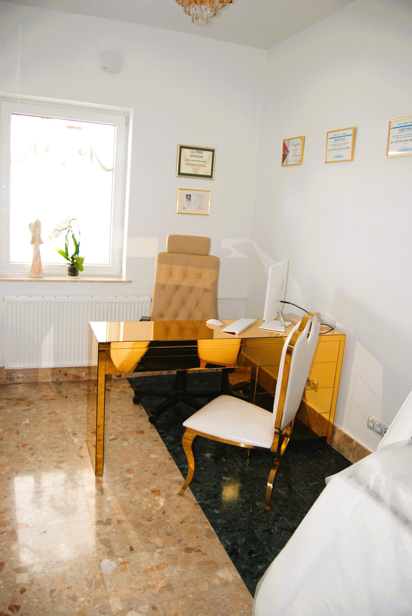 Jasne wnętrze gabinetu z biurkiem w kolorze złota, białym krzesłem i dyplomami na ścianie, widok na okno z rośliną i figurką.