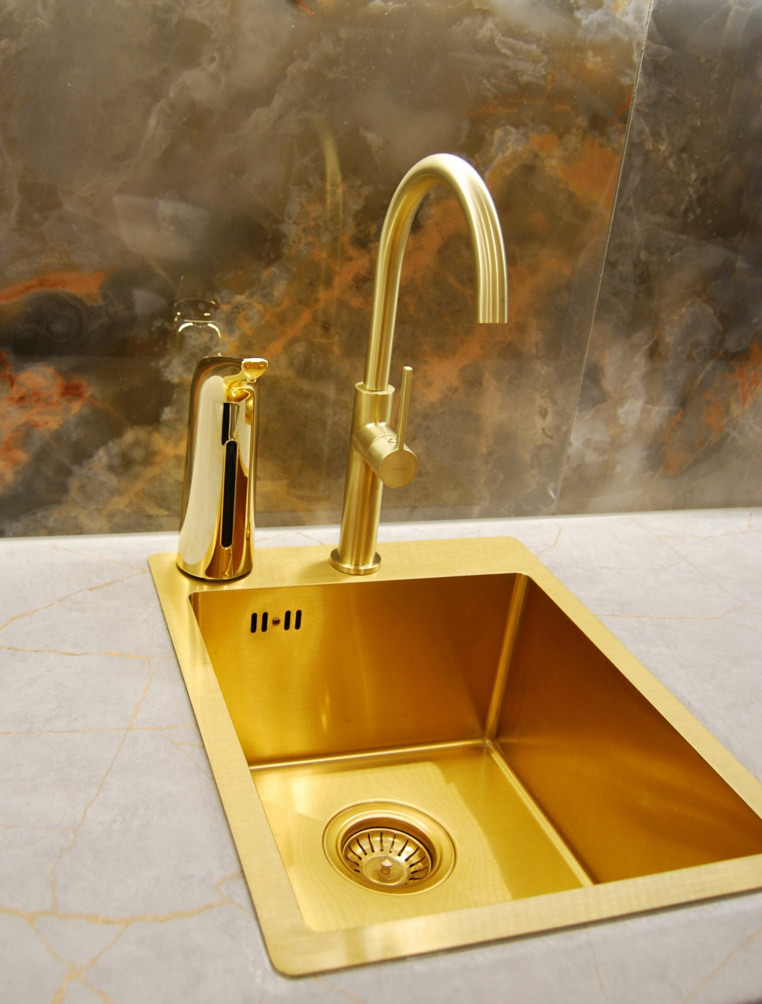 Złota umywalka z baterią i dozownikiem mydła na blacie z marmurowym wzorem, tło stanowi ściana z podobnym wzorem.