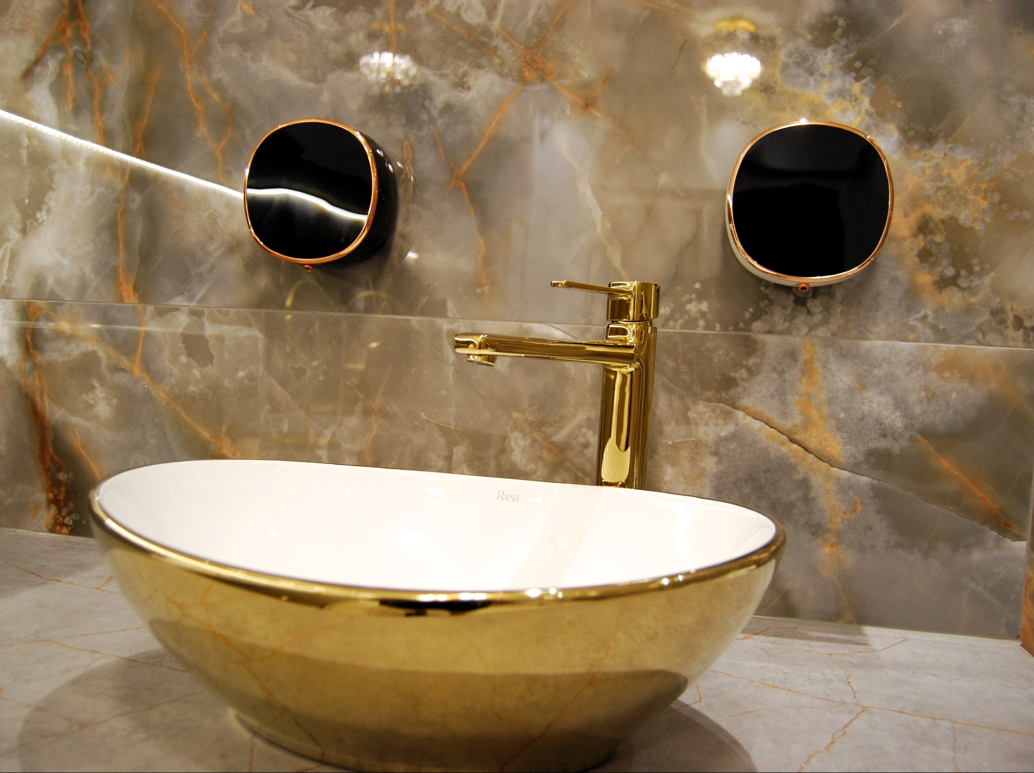 Biała umywalka ze złotym zdobieniem, bateria w kolorze złota i okrągłe, czarne lustra na ścianie z marmurowym wzorem.