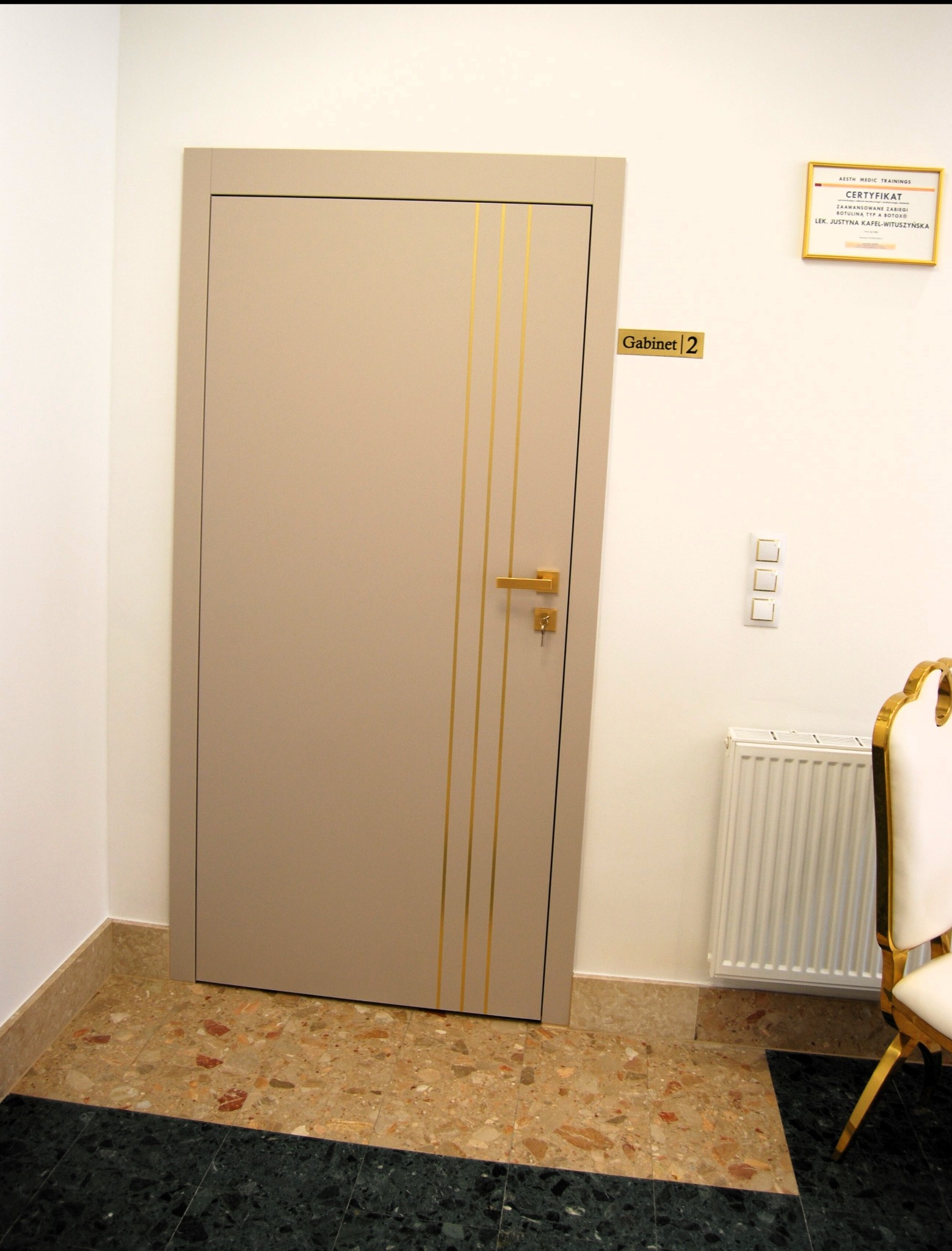 Beżowe drzwi z trzema złotymi paskami do gabinetu numer 2, na ścianie certyfikat i biały grzejnik, fragment złotego krzesła, marmurowa podłoga w dwóch odcieniach.