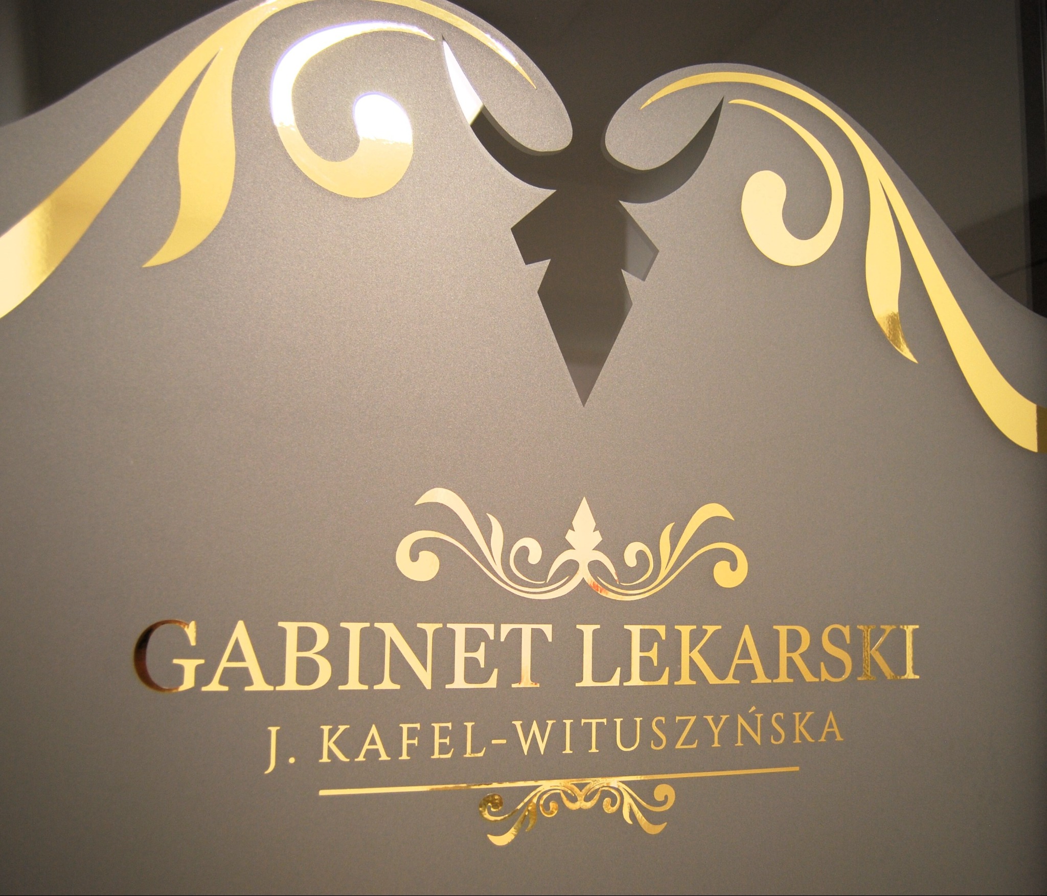Złoty napis 'GABINET LEKARSKI J. KAFEL-WITUSZYŃSKA' na szarej, matowej powierzchni z dekoracyjnymi ornamentami.