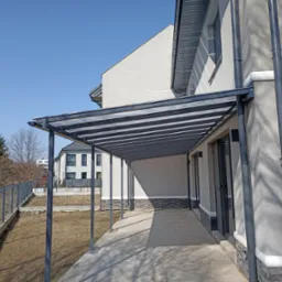 Nowoczesna pergola metalowa z przezroczystym zadaszeniem, przylegająca do ściany budynku, z widocznym systemem odprowadzania wody i metalowym ogrodzeniem w tle.