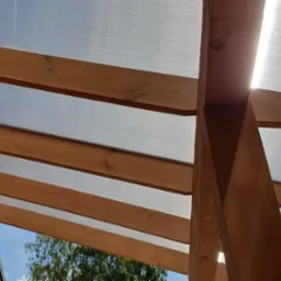 Drewniana konstrukcja altany z przezroczystym poliwęglanowym dachem, widok od dołu w słoneczny dzień.