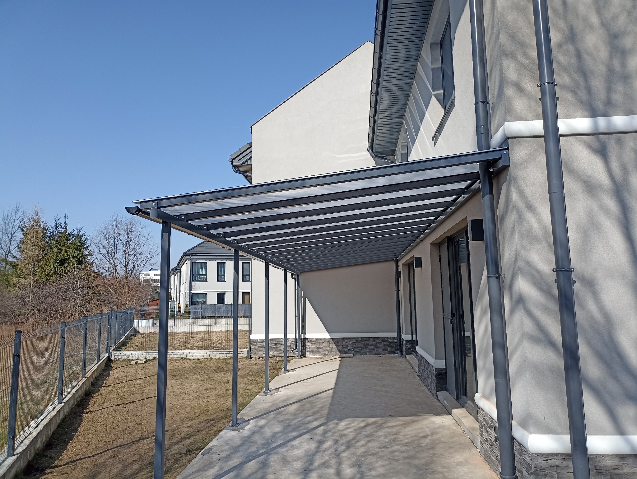 Nowoczesna pergola metalowa z przezroczystym zadaszeniem, przylegająca do ściany budynku, z widocznym systemem odprowadzania wody i metalowym ogrodzeniem w tle.
