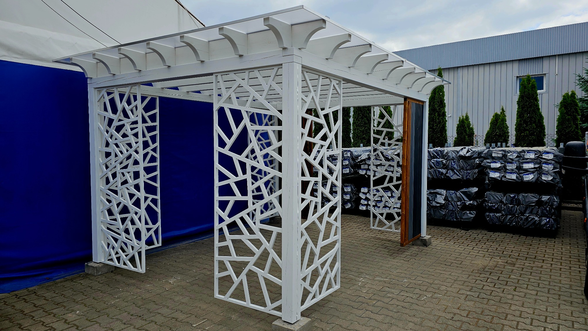 Biała pergola z ażurowymi ścianami o geometrycznym wzorze, na tle niebieskiej plandeki i zapakowanych materiałów budowlanych.