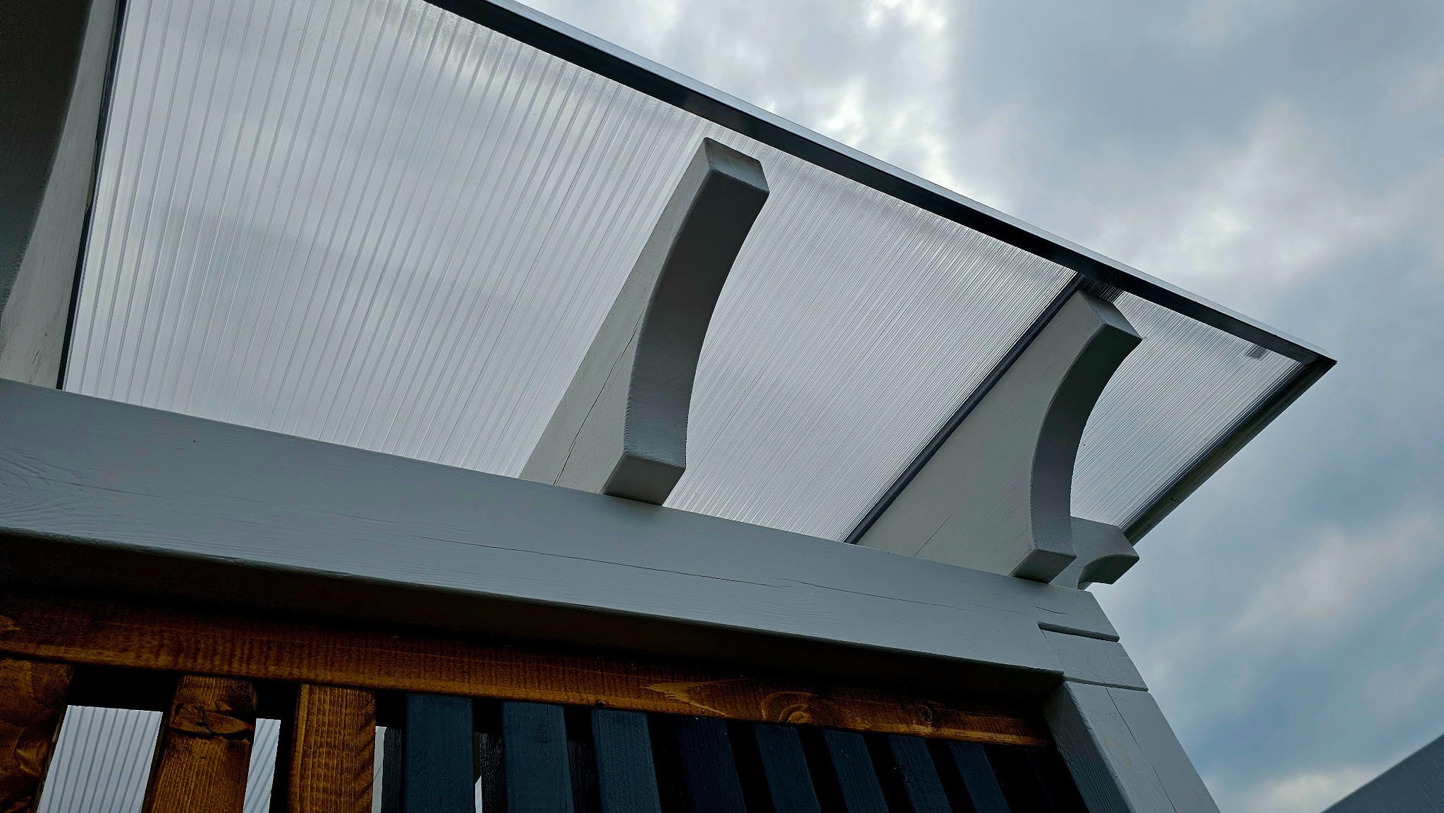 Fragment konstrukcji z przezroczystym zadaszeniem, wspartej na drewnianych elementach pomalowanych na szaro i brązowo, widziana z dołu na tle pochmurnego nieba.