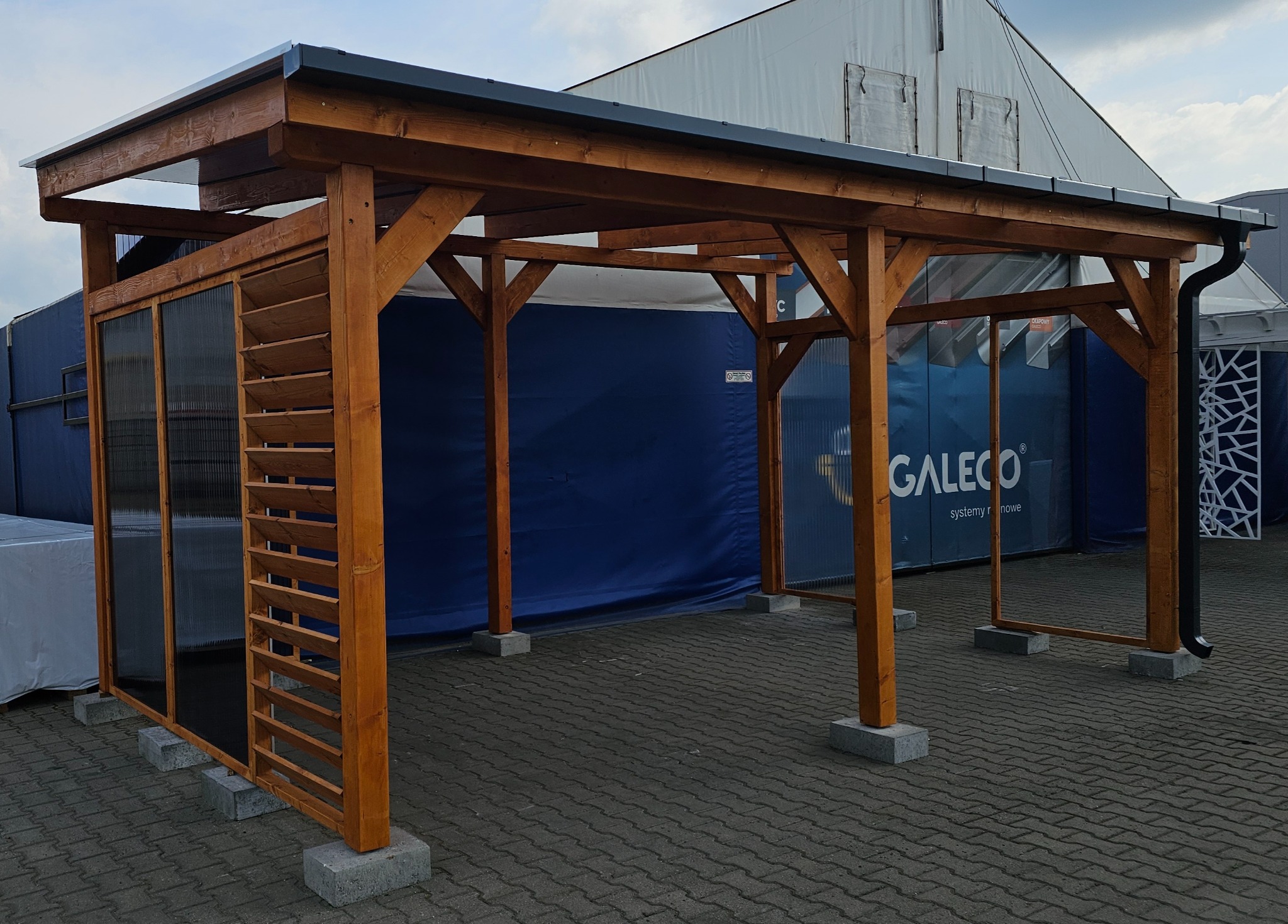 Drewniana konstrukcja z zadaszeniem, częściowo osłonięta lamelami i przezroczystymi panelami, ustawiona na betonowych bloczkach, na tle namiotu z reklamą.