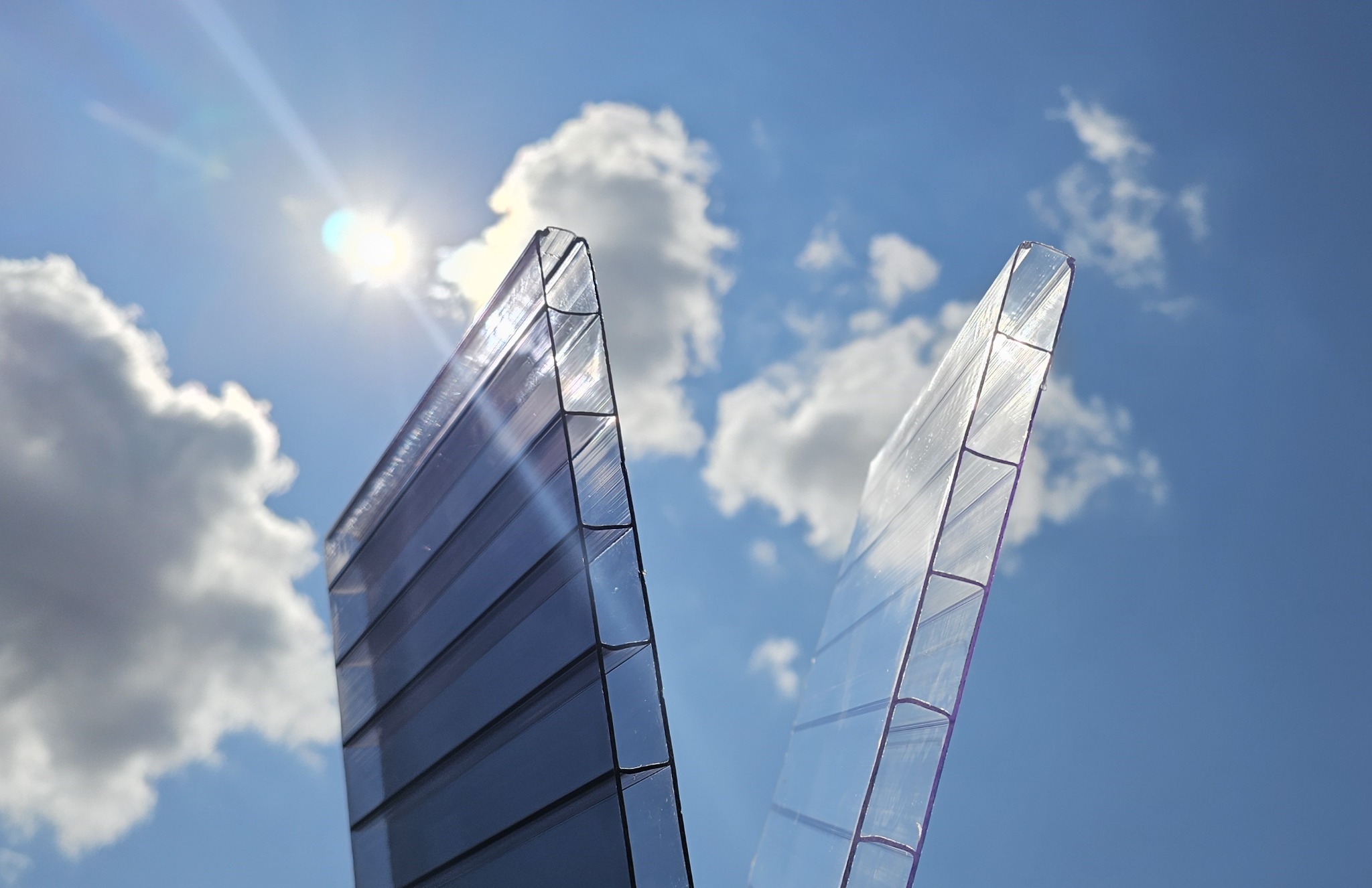 Dwa fragmenty przezroczystego poliwęglanu komorowego na tle błękitnego nieba z chmurami i słońcem, ukazujące strukturę materiału.