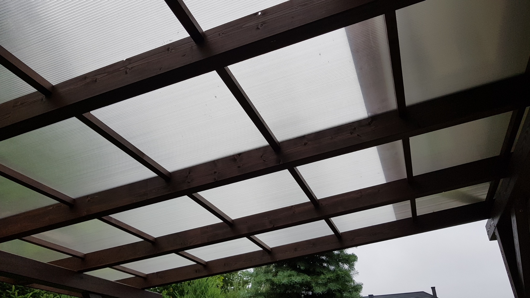 Drewniana konstrukcja z przezroczystym zadaszeniem, widok z dołu, ukazujący regularny układ belek i paneli przepuszczających światło.