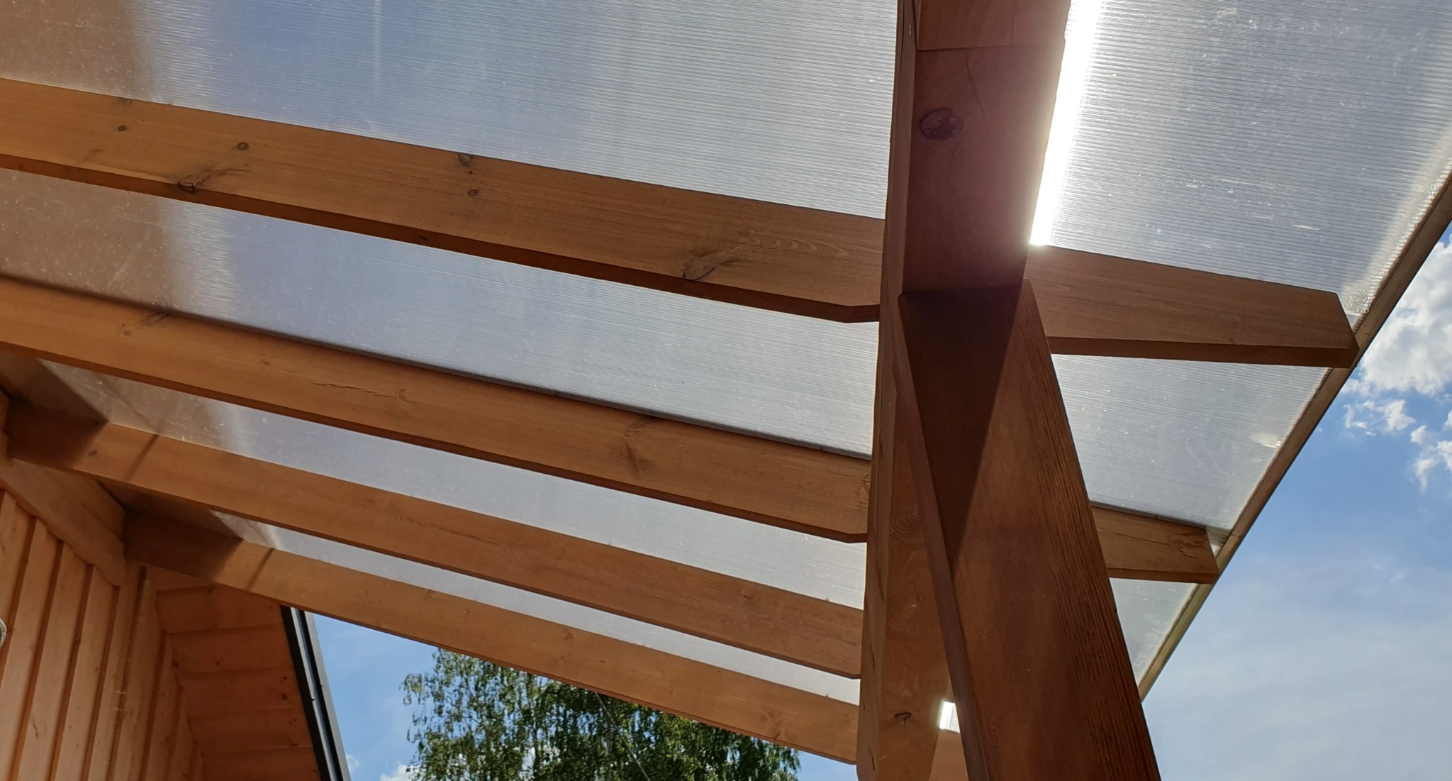 Drewniana konstrukcja altany z przezroczystym poliwęglanowym dachem, widok od dołu w słoneczny dzień.