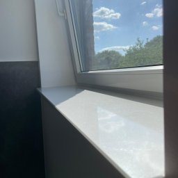 PIOTR ROZMYSŁOWSKI - Biały parapet okienny z blatem imitującym marmur, widok na zewnątrz przez okno dachowe z moskitierą, fragment ciemnej ściany obok.
