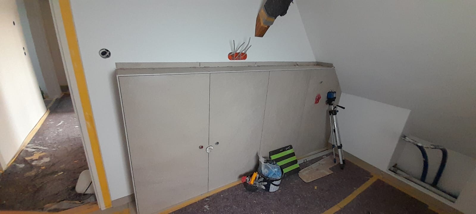 Poddasze w trakcie adaptacji: zabudowa z płyt kartonowo-gipsowych z wywierconymi otworami na zamki, widoczne przewody elektryczne, poziomica laserowa na statywie oraz narzędzia w torbie.