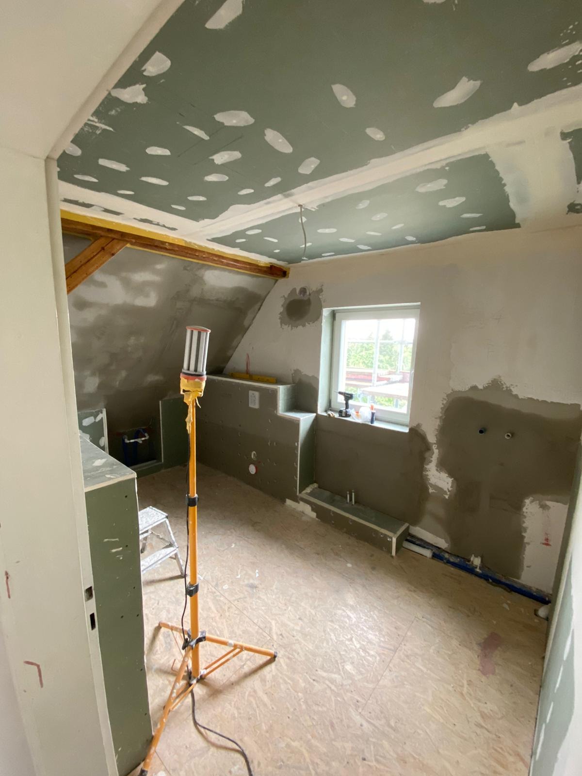 Poddasze w trakcie prac wykończeniowych: ściany i sufit pokryte szpachlą, widoczne instalacje, lampa budowlana na statywie, okno z widokiem na zieleń.