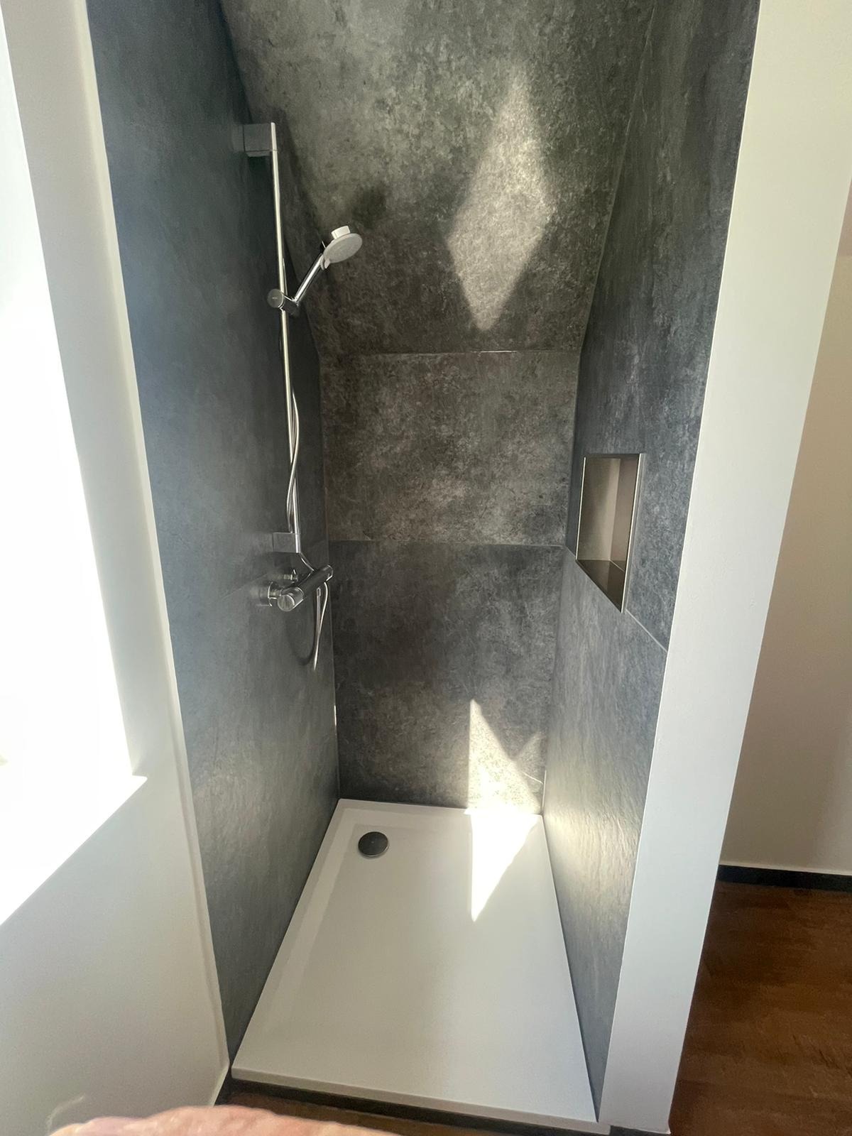 Nowoczesna kabina prysznicowa z szarymi ścianami imitującymi beton, biały brodzik i wnęka w ścianie, oświetlona naturalnym światłem.