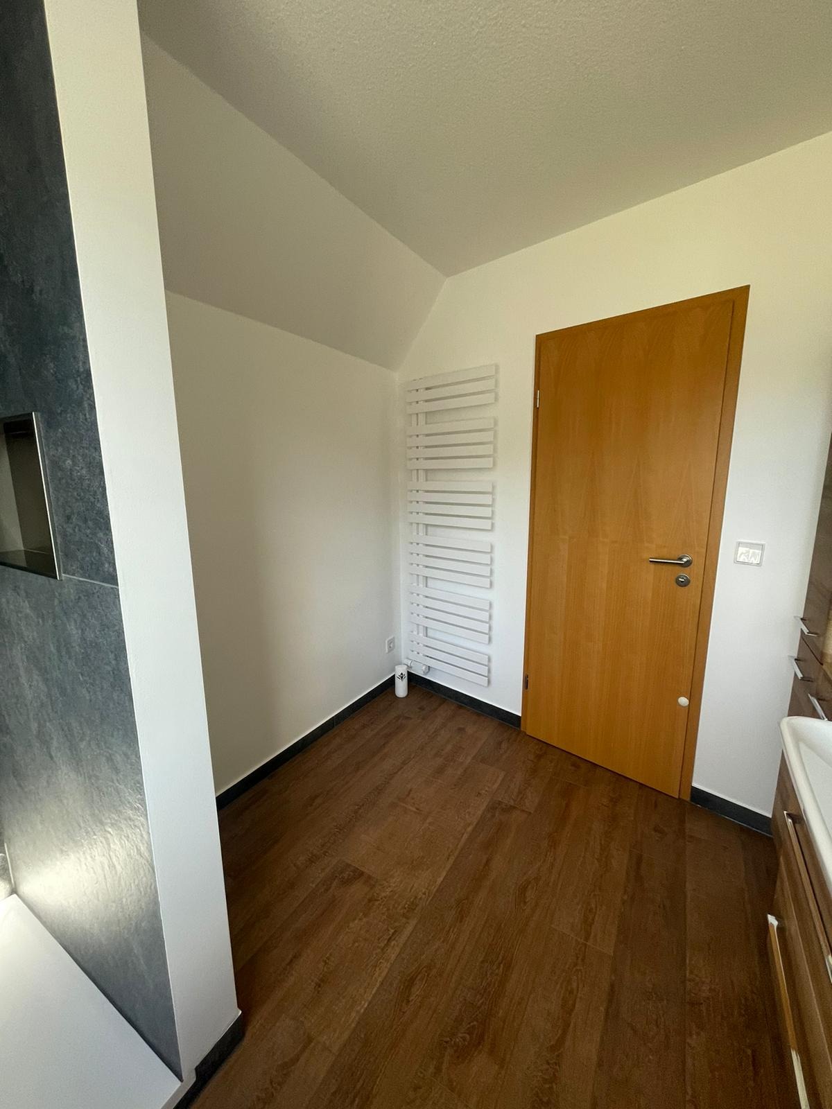 Nowoczesna łazienka z drewnopodobną podłogą, grzejnikiem drabinkowym i drzwiami z jasnego drewna, widok na narożnik z szarą płytką.