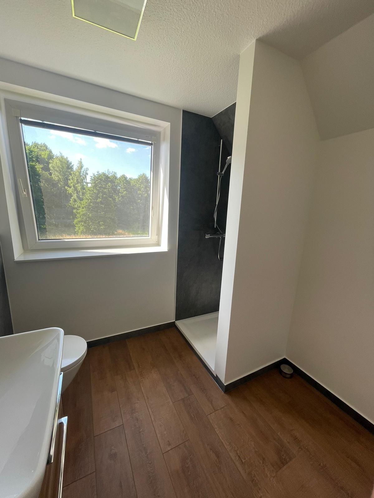 Jasna łazienka z drewnopodobną podłogą, kabiną prysznicową z ciemną ścianą i dużym oknem z widokiem na zieleń.