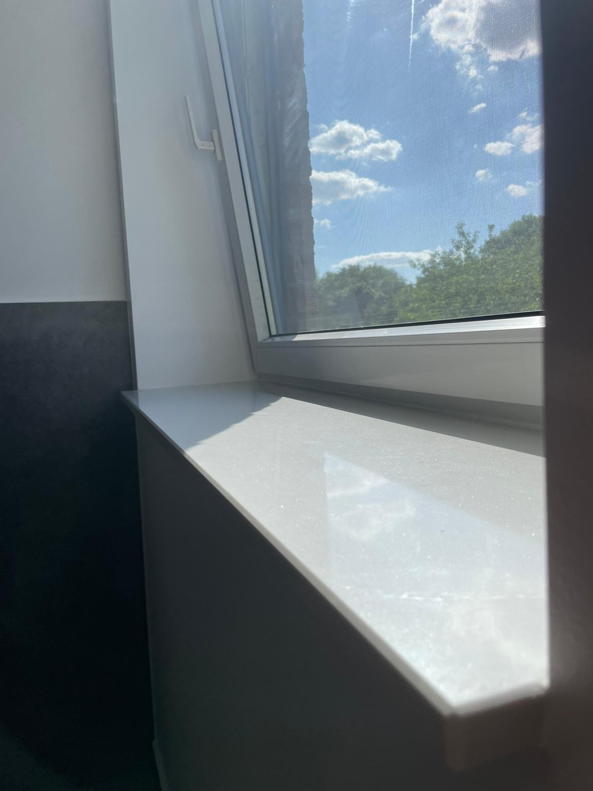 Biały parapet okienny z blatem imitującym marmur, widok na zewnątrz przez okno dachowe z moskitierą, fragment ciemnej ściany obok.