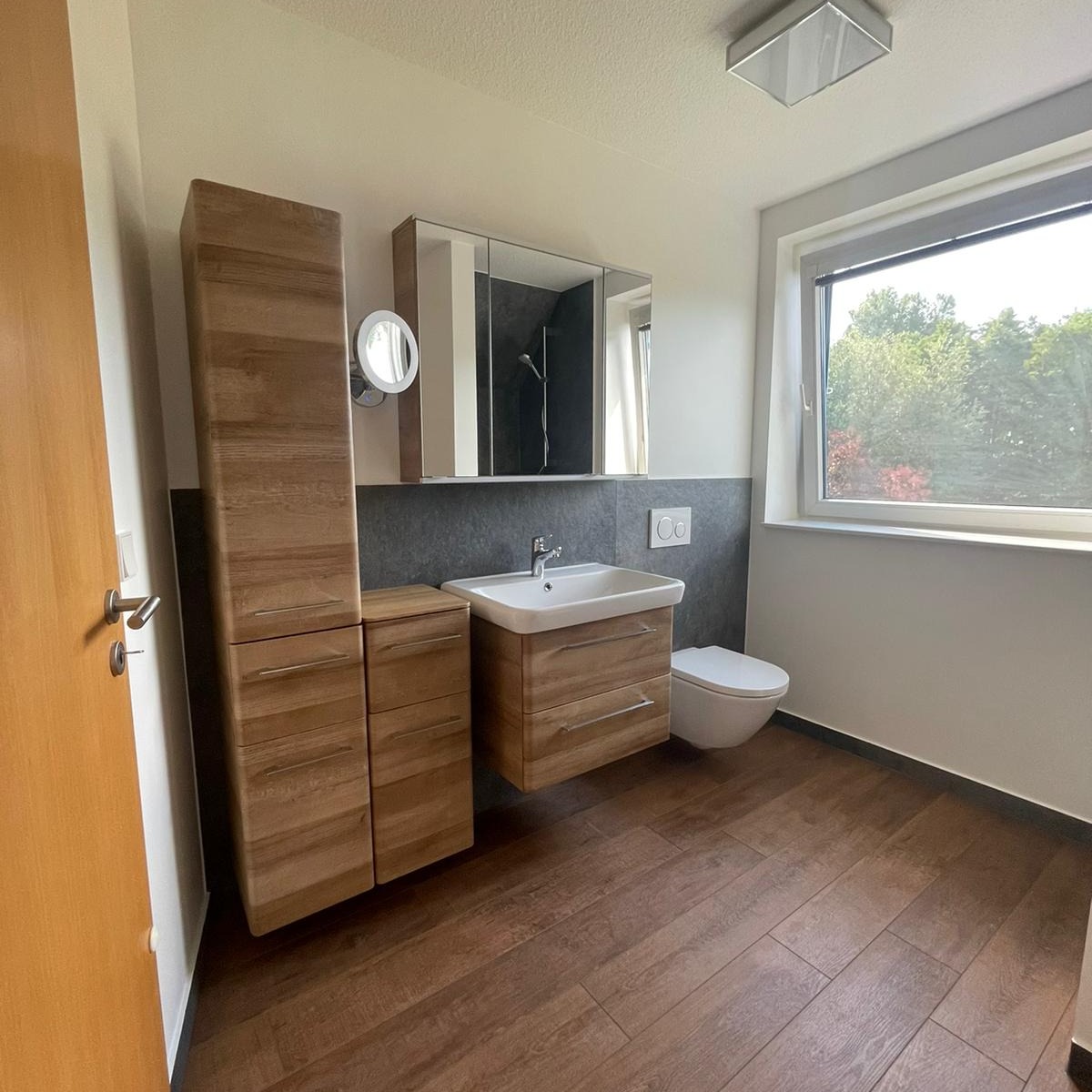 Jasna łazienka z drewnianymi meblami: wysoka szafka, szafka pod umywalką z szufladami, lustro z oświetleniem, biała umywalka i toaleta, brązowa podłoga imitująca drewno, szara ściana za umywalką.