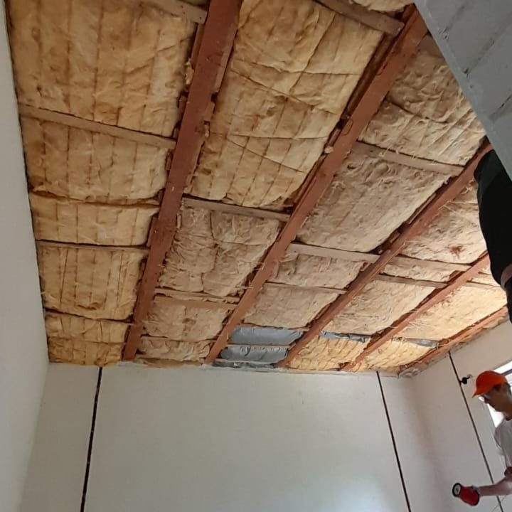 Montaż izolacji termicznej z wełny mineralnej na drewnianej konstrukcji sufitu w Ostrowcu Świętokrzyskim, widoczny fragment ściany i osoba w pomarańczowej czapce.