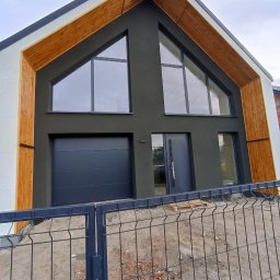 Łukasz Buczma - Nowoczesny dom jednorodzinny z grafitową elewacją, drewnianymi akcentami i dużymi oknami trójkątnymi, widziany zza metalowego ogrodzenia.