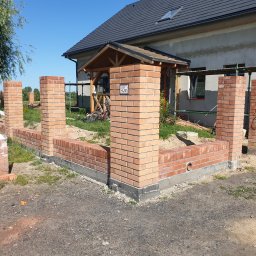 Łukasz Buczma - Ceglany murek z filarami w trakcie budowy przed domem z szarą elewacją i czarnym dachem. Widoczne rusztowania i materiały budowlane.