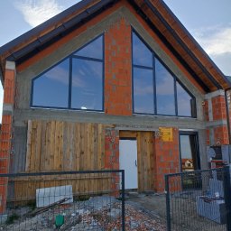 Łukasz Buczma - Surowy stan domu jednorodzinnego z czerwonej cegły z zamontowanymi oknami o nietypowym kształcie, zabezpieczony drewnianą konstrukcją, widok zza metalowego ogrodzenia.