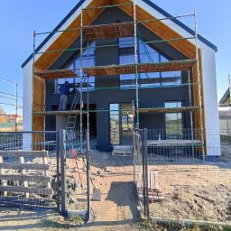 Łukasz Buczma - Nowoczesny dom w trakcie malowania elewacji na szaro, z drewnianymi elementami wykończeniowymi. Widoczne rusztowania i pracownik na drabinie. Błękitne niebo w tle.