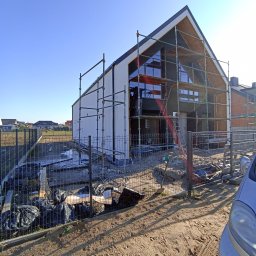 Łukasz Buczma - Dom jednorodzinny w trakcie ocieplania, widoczne rusztowania i częściowo otynkowana elewacja. W tle błękitne niebo i inne domy.