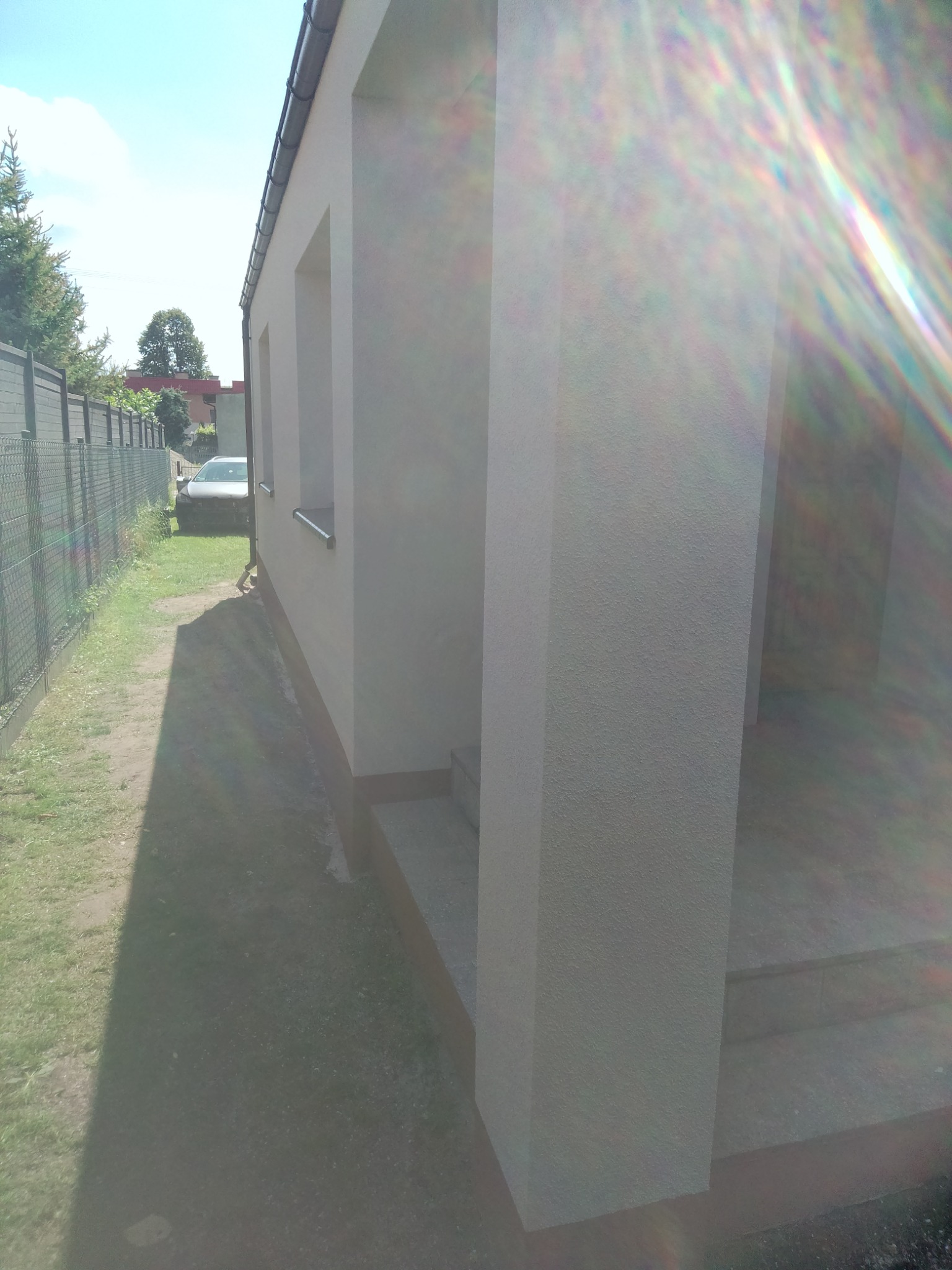 Świeżo otynkowana elewacja domu jednorodzinnego z widocznym fragmentem ogrodzenia i samochodu w tle, silne refleksy słoneczne.