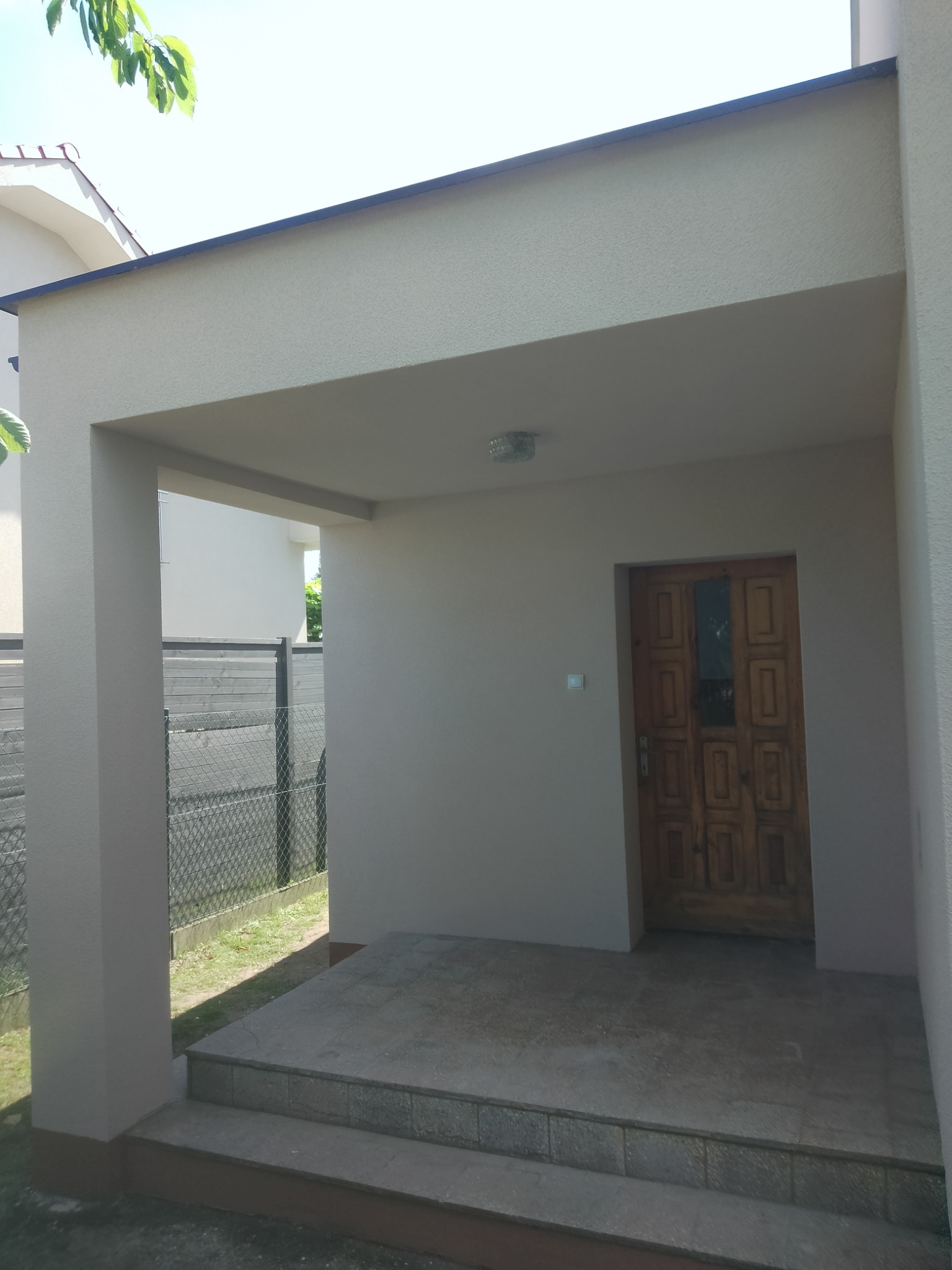 Zadaszone wejście do domu z drewnianymi drzwiami, widoczne trzy kamienne stopnie i oświetlenie sufitowe.